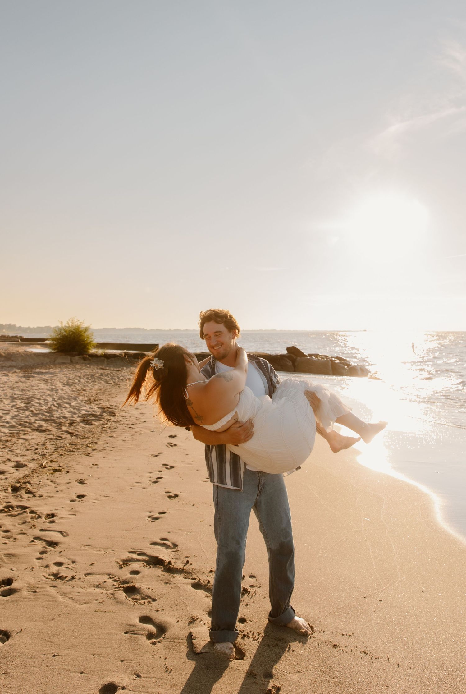 A man lifts a woman in a white dress on a sunny beach at golden hour, waves and sandy shore in the background.