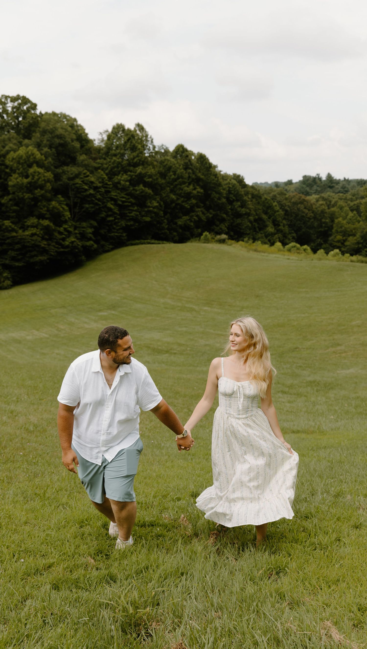 A couple holds hands while joyfully running together through a lush green cuyahoga hillside meadow on an overcast summer day.