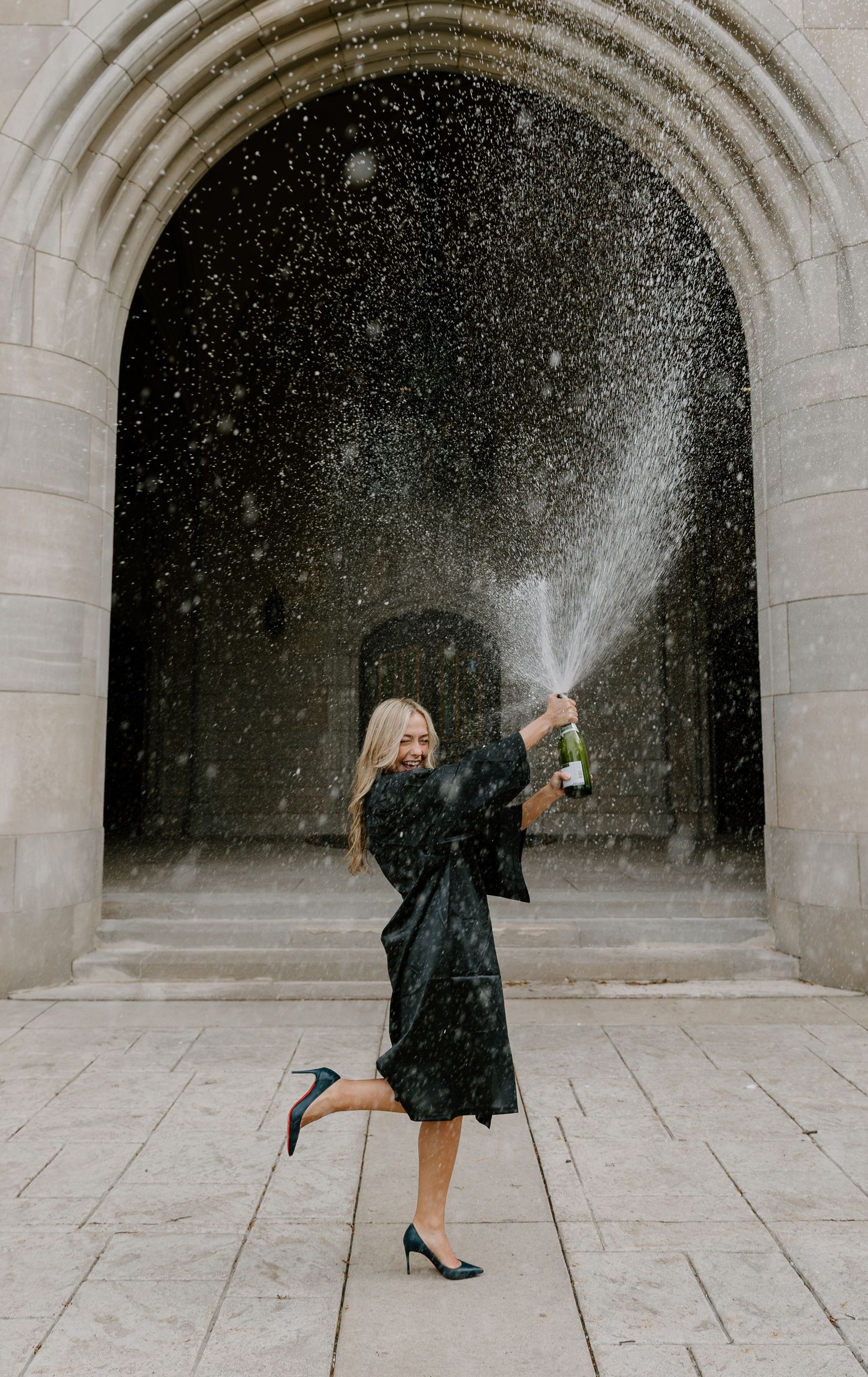 Woman in black dress joyfully spraying champagne under a stone arch, celebrating a milestone occasion.