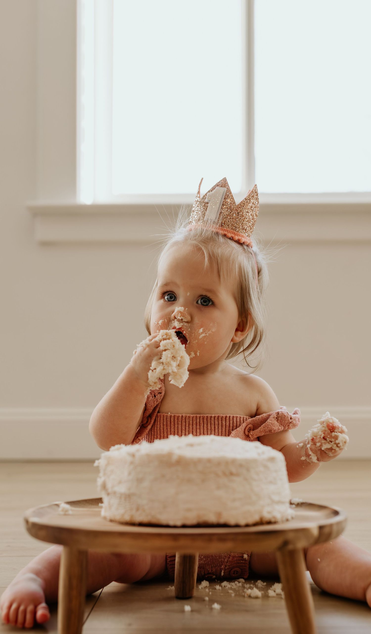 Toddler wearing crown enthusiastically eating white cake from wooden table.