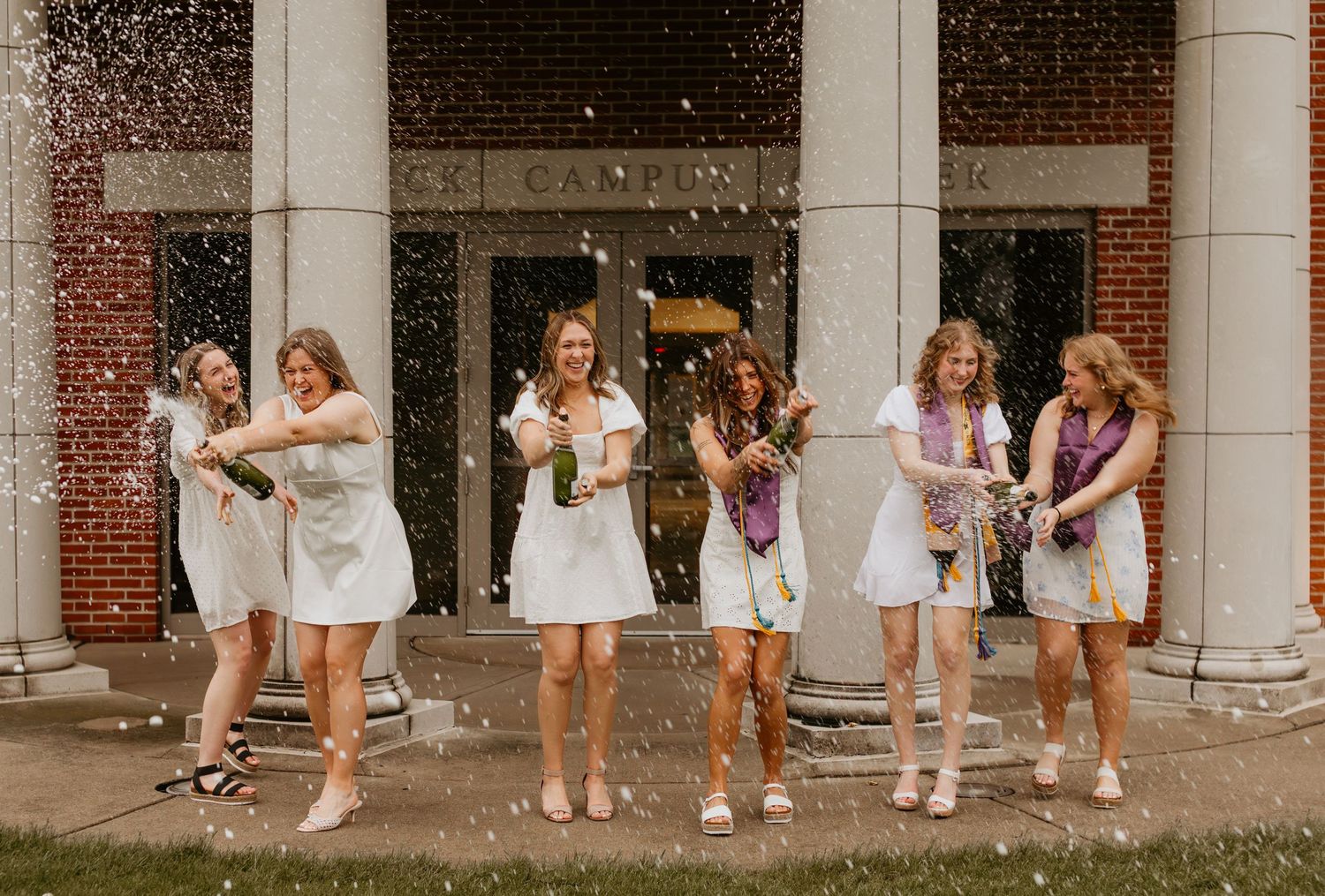 A group in white dresses and sneakers dance joyfully together under a columned building entrance.