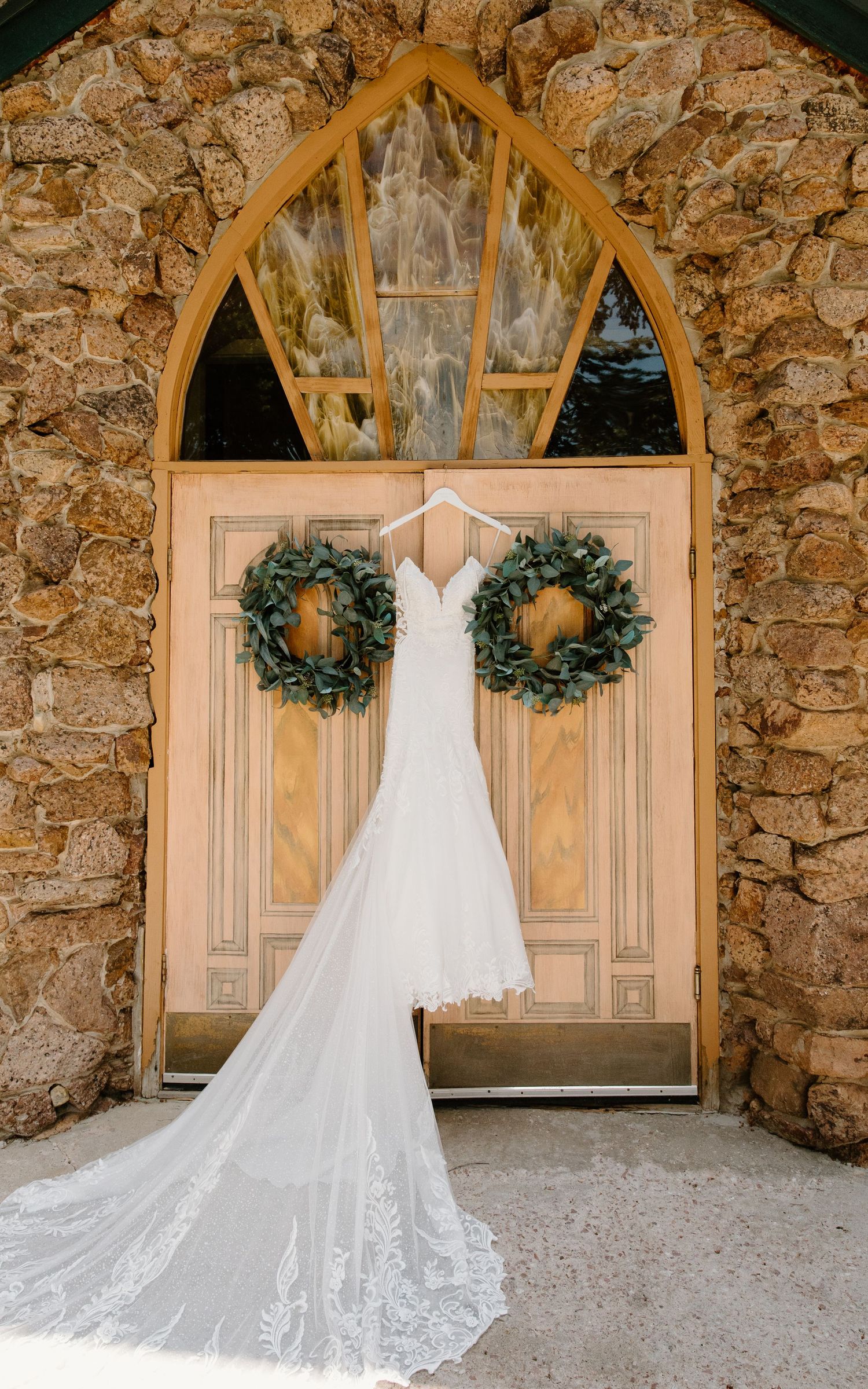 A stunning white lace wedding dress hangs elegantly against rustic stone walls with decorative wreaths and arched windows.
