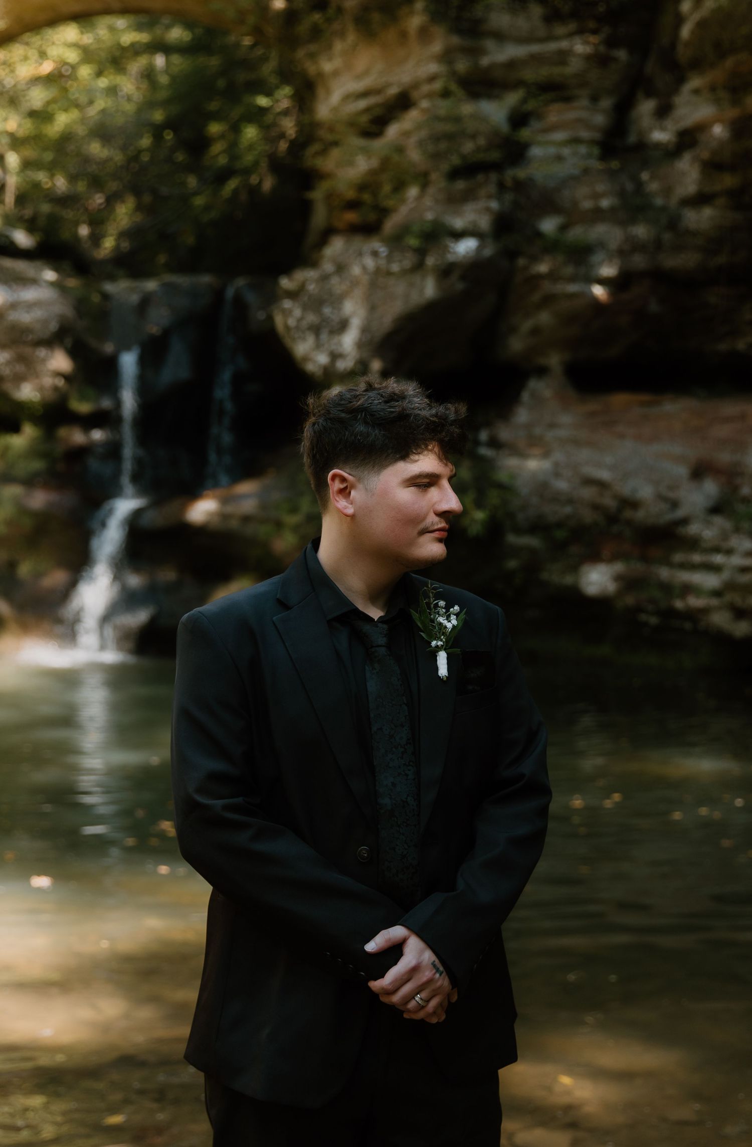 A groom in a black suit with boutonniere stands before a serene waterfall and rocky backdrop.