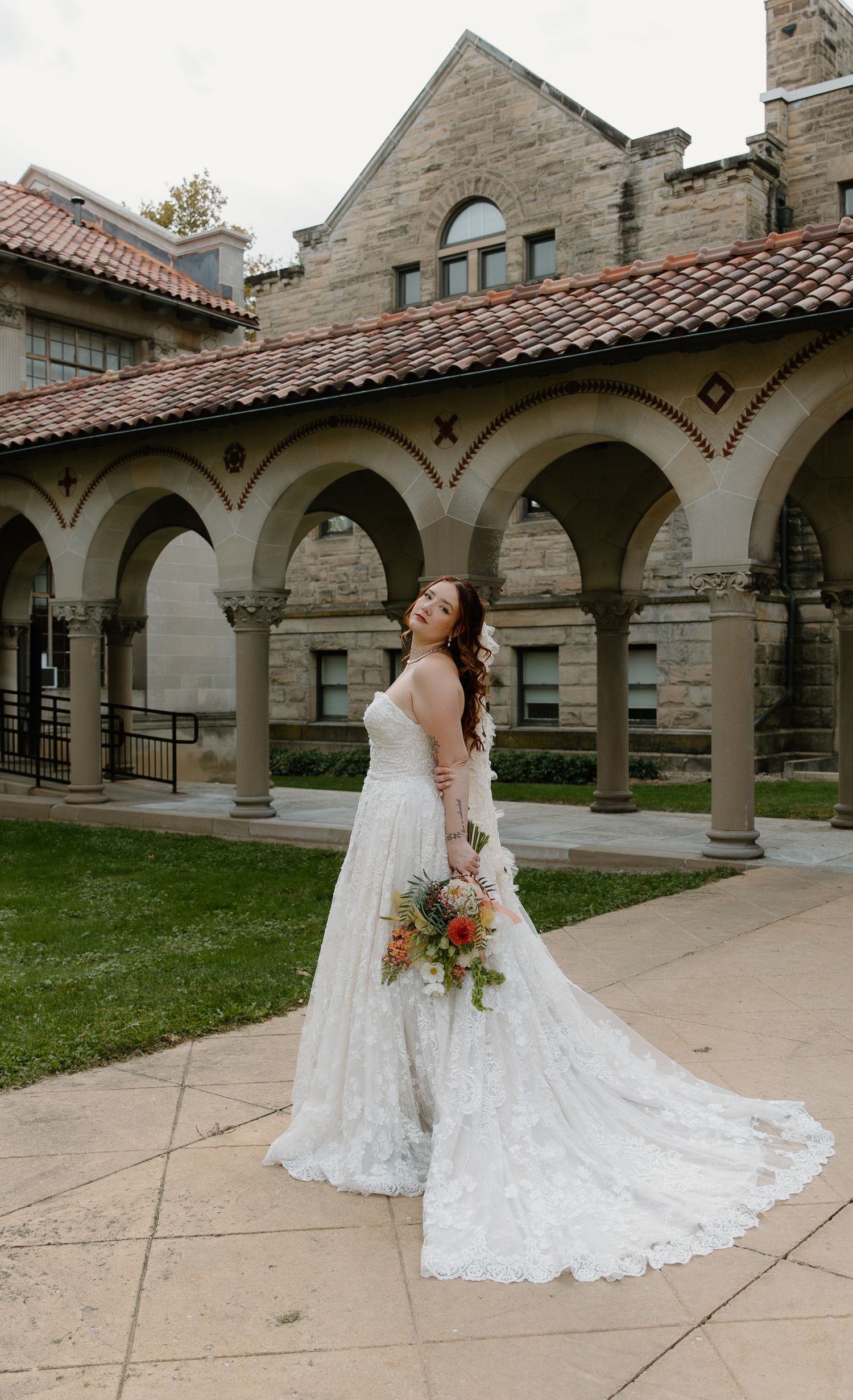 Bride in lace wedding gown holding colorful bouquet poses before stone arched colonnade building.