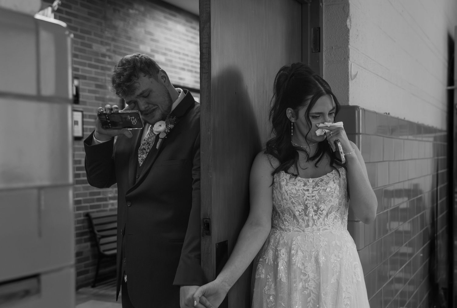 Black and white photo of emotional bride and groom holding hands, each wiping tears on their wedding day.