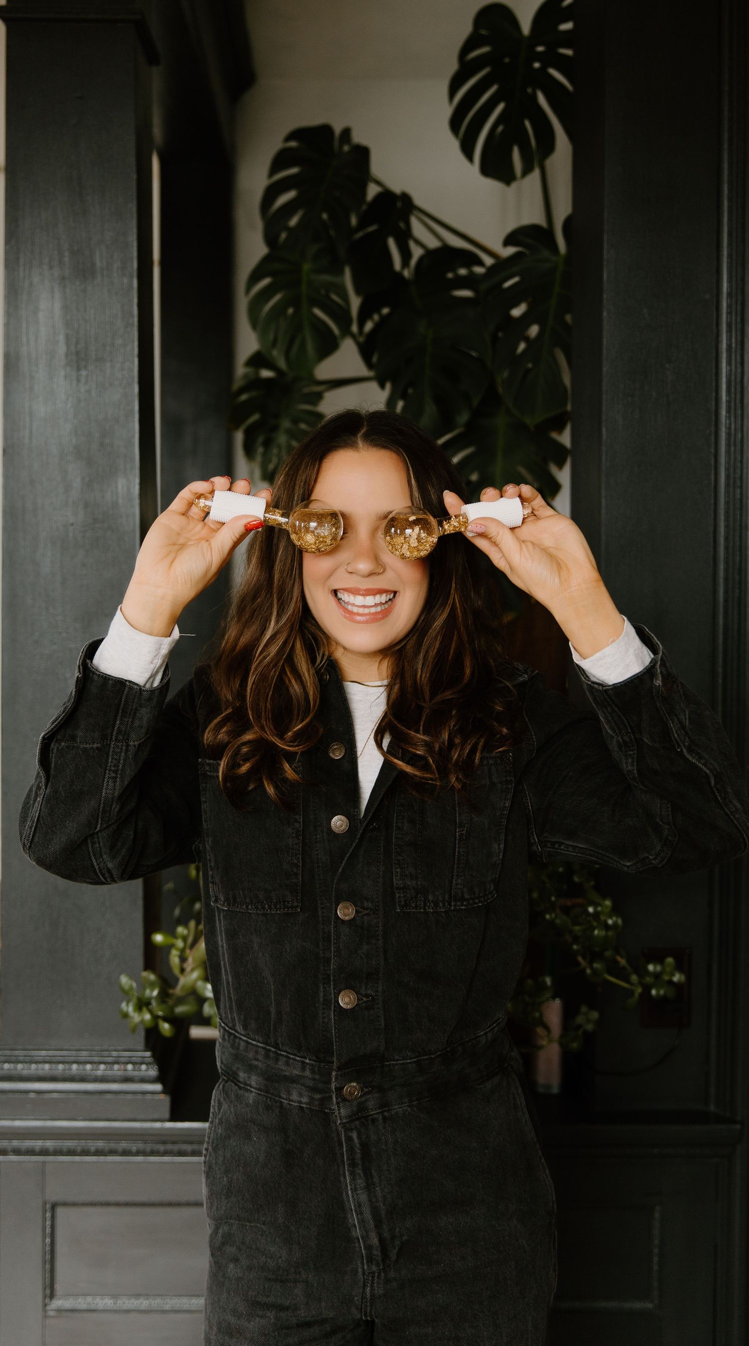 Smiling woman in black denim jacket holding gold sunglasses, posing in front of monstera plant.