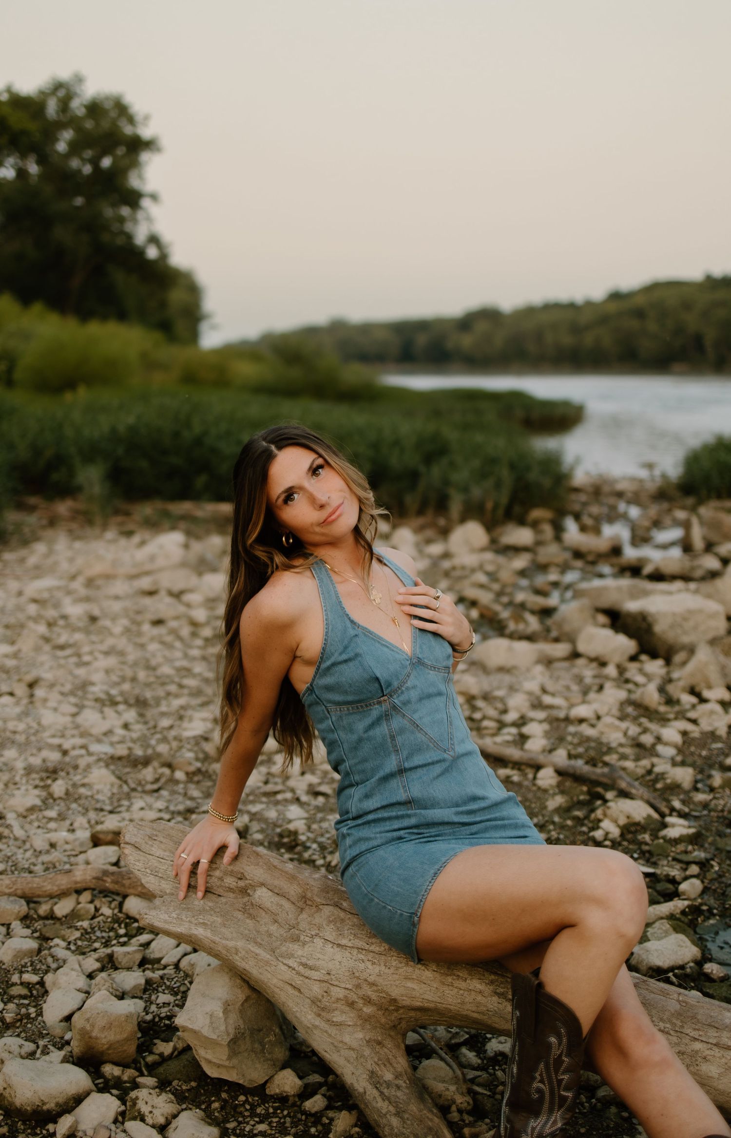 A woman in a blue mini dress poses on driftwood by a rocky riverbank with lush green trees in the background.