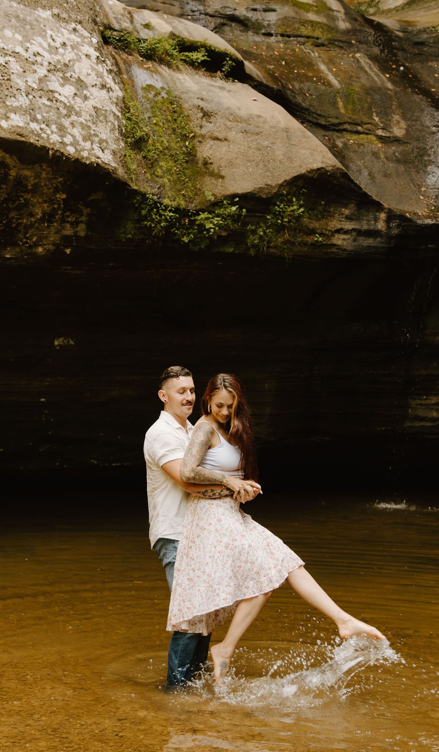 Couple in white clothing playfully splashing in shallow brown water beneath a rocky overhang during a romantic photoshoot.