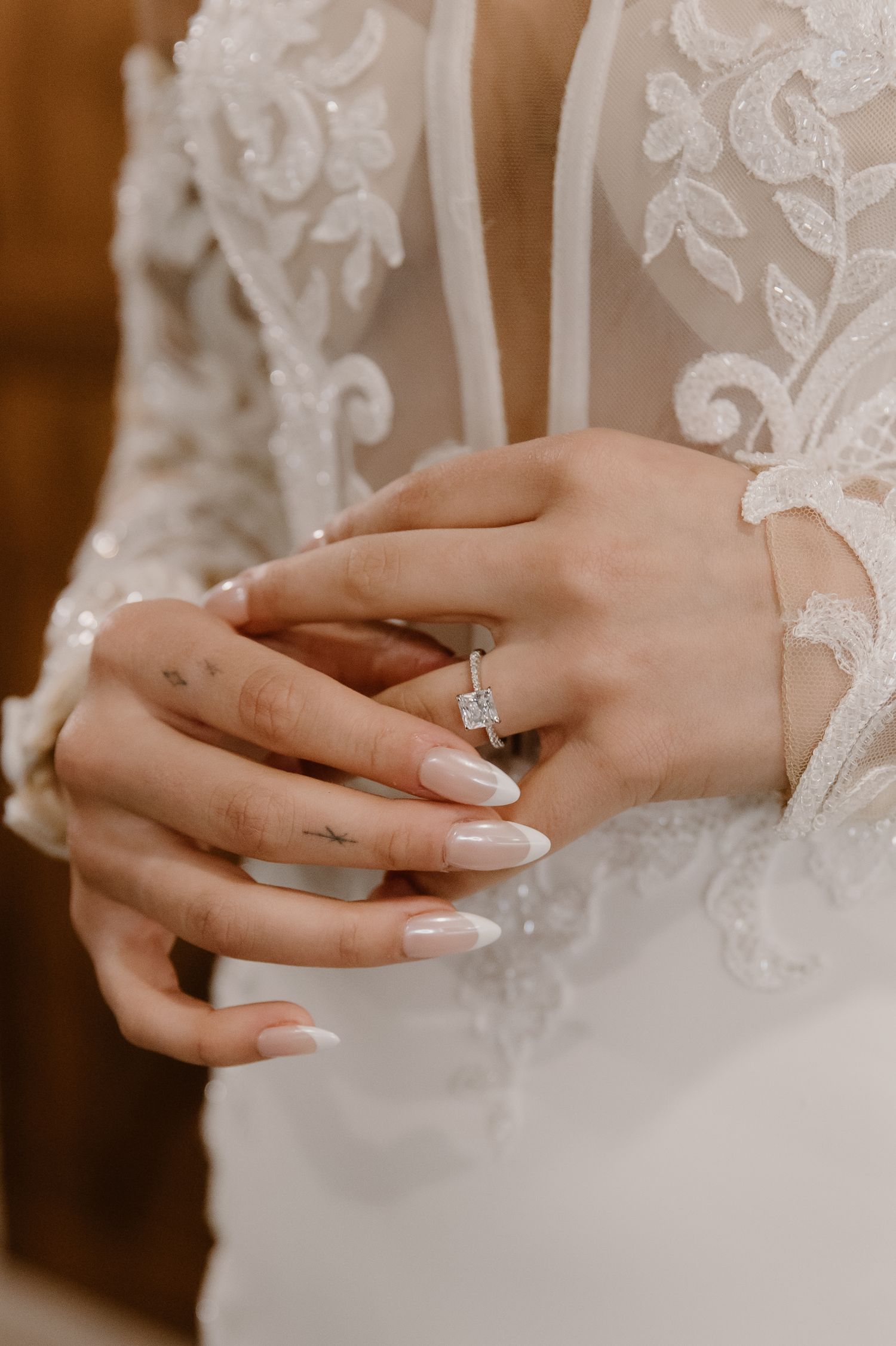 Bride in lace wedding gown clasping hands showing elegant diamond engagement ring with almond-shaped French manicure nails.