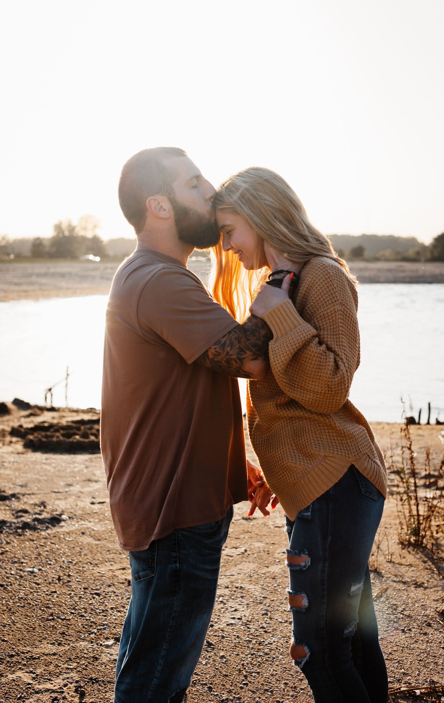 Couple sharing a tender moment at sunset by a lake wearing cozy sweaters.