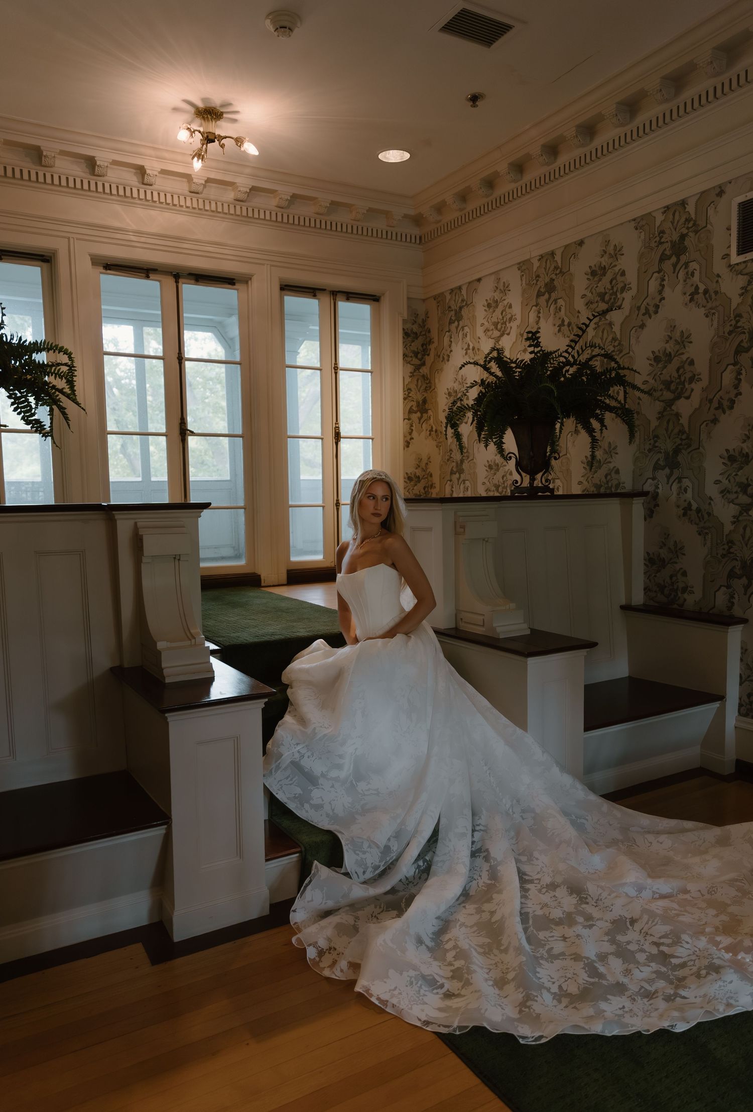 Elegant bride in a strapless lace ball gown seated in a vintage room with floral wallpaper.