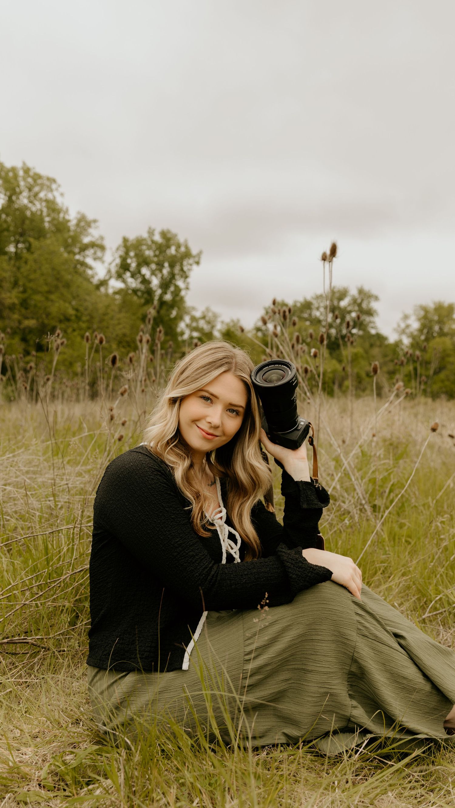 A person in black clothing relaxes in a grassy field on an overcast day with their black cat companion.