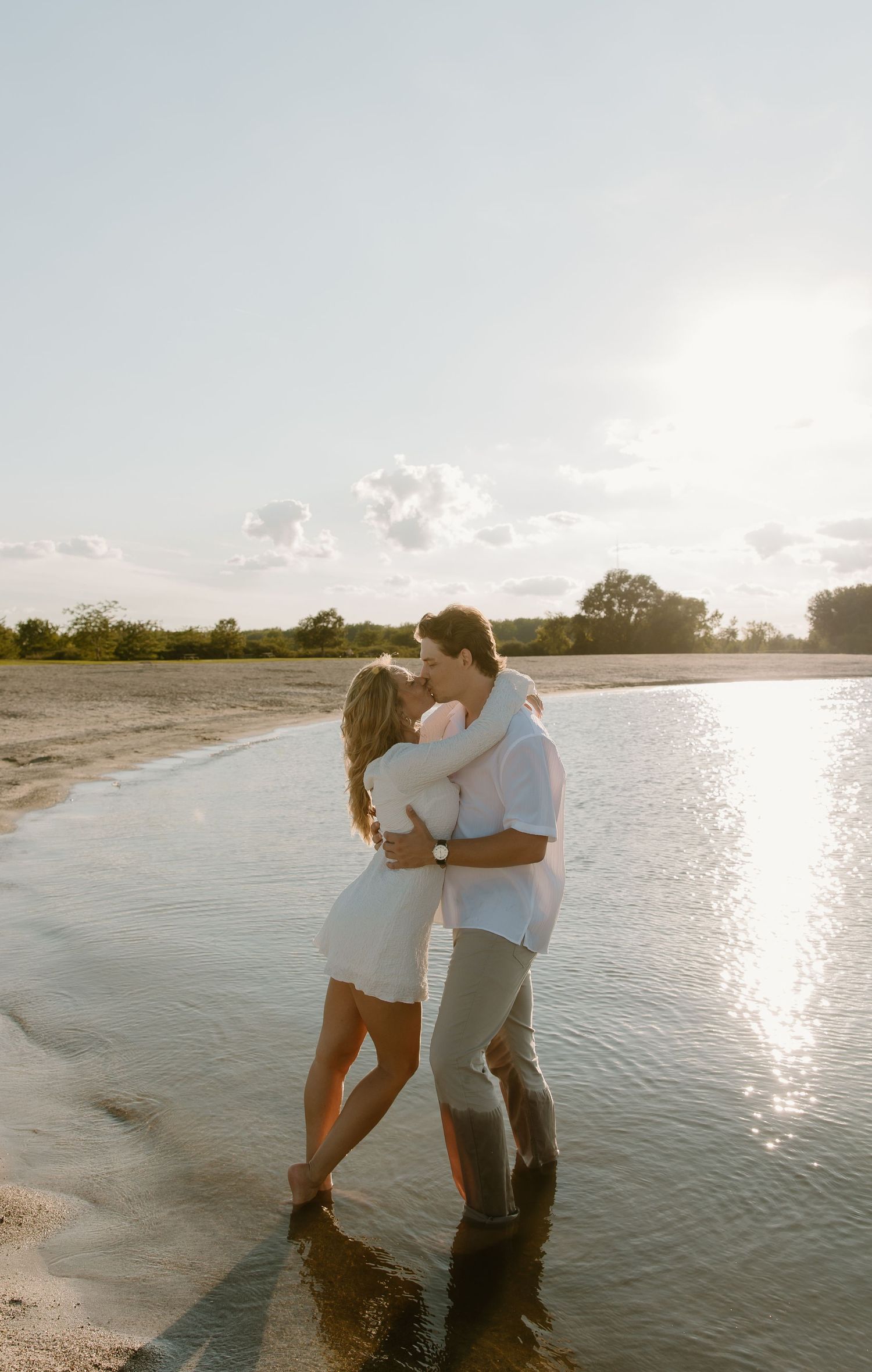 Couple kissing while standing in shallow water on a sandy beach at golden hour sunset.