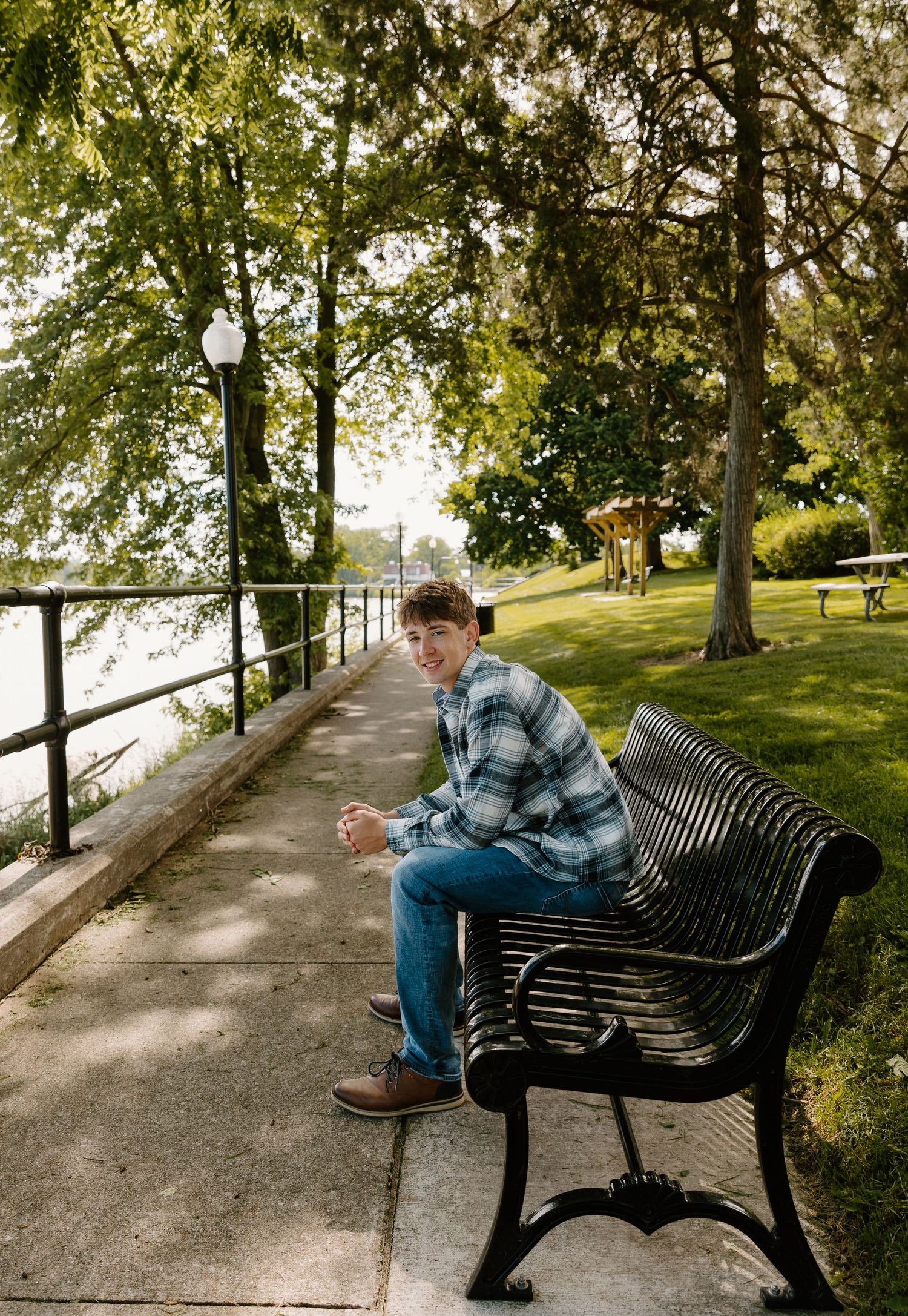 Young man in plaid shirt sitting on a park bench by a waterfront path surrounded by lush green trees.