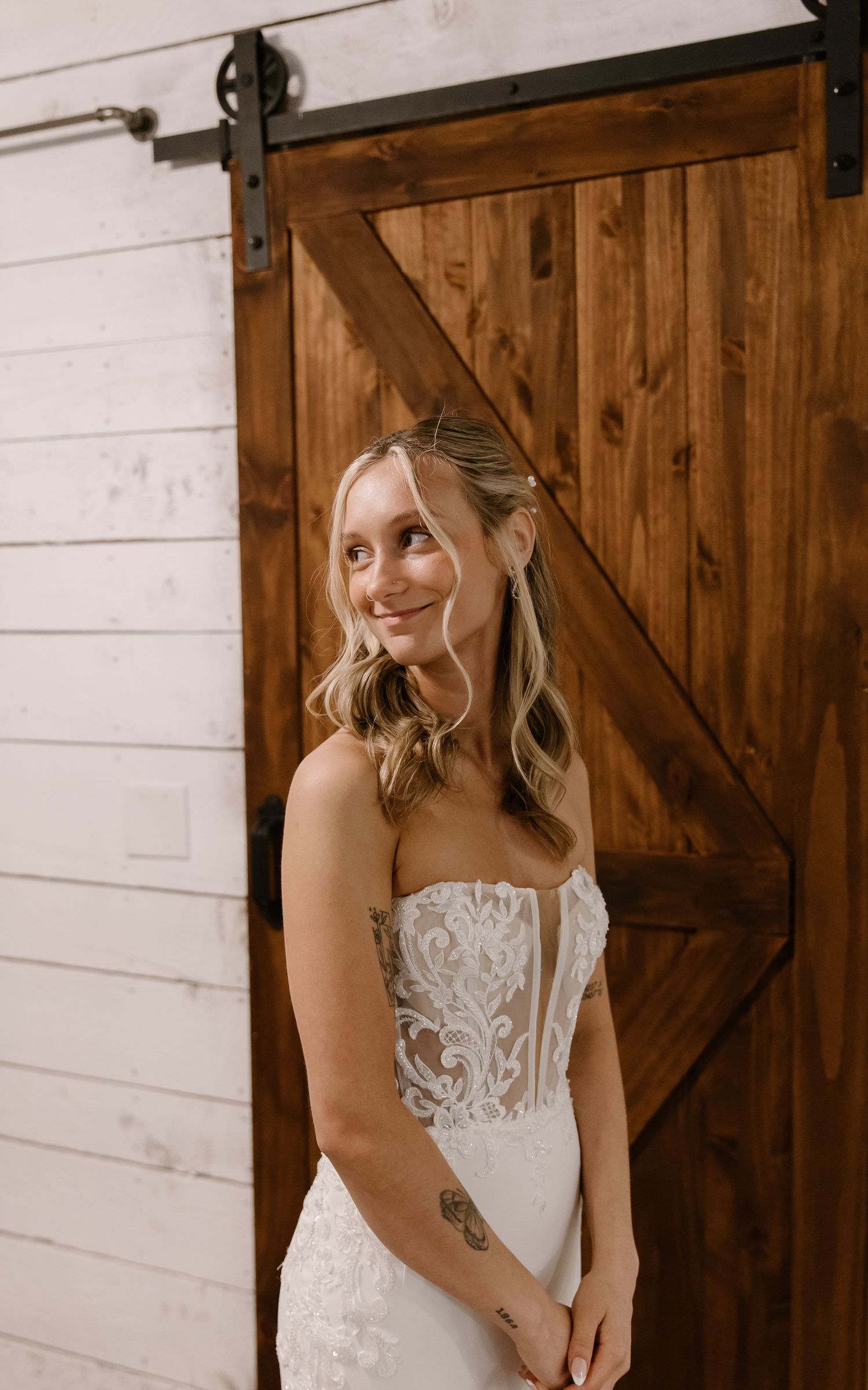 Smiling bride in strapless lace wedding gown standing in front of rustic wooden barn door.