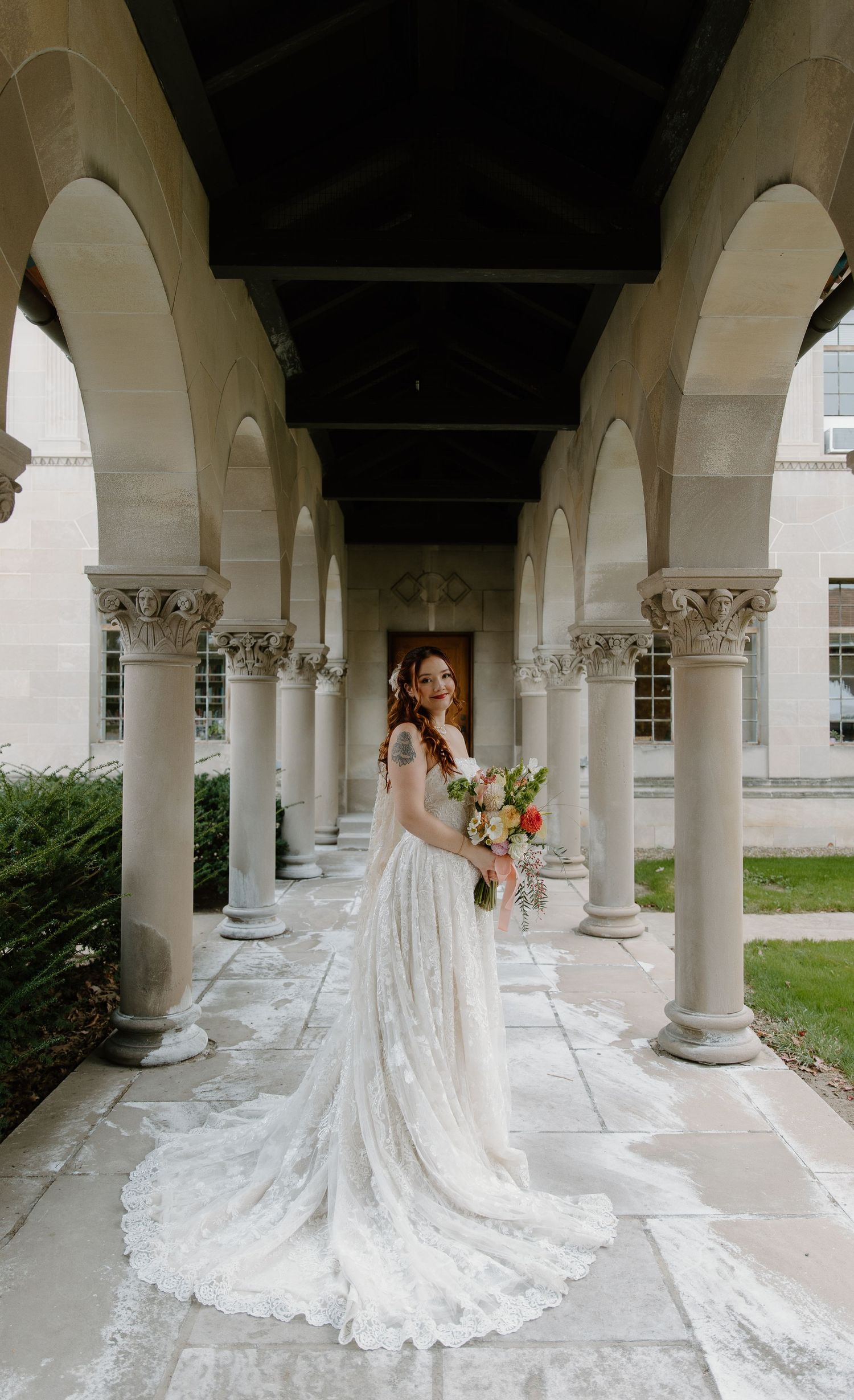 Bride in lace gown with long train poses among stone arched columns holding colorful bouquet at elegant venue.