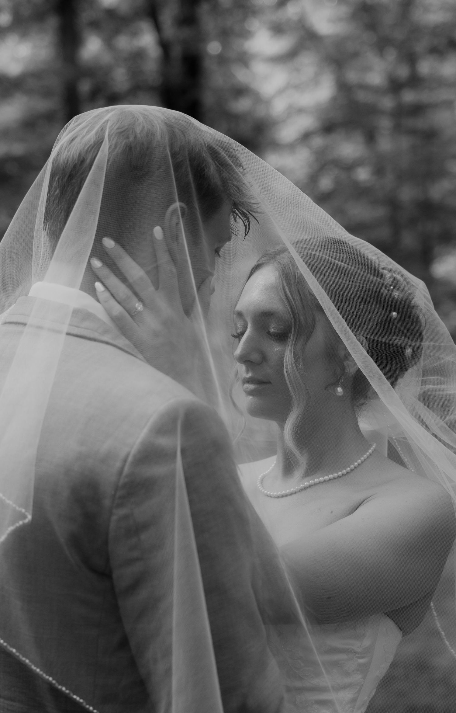 Black and white wedding photo of bride and groom sharing intimate moment under flowing veil outdoors.
