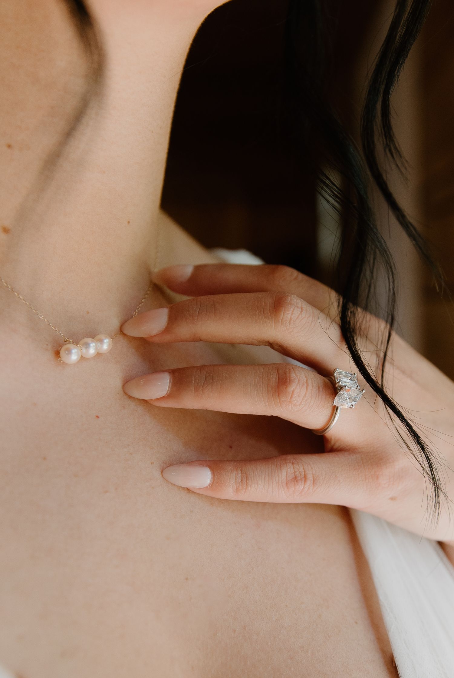 Close-up of a bride wearing a diamond engagement ring and pearl necklace, with manicured nails.