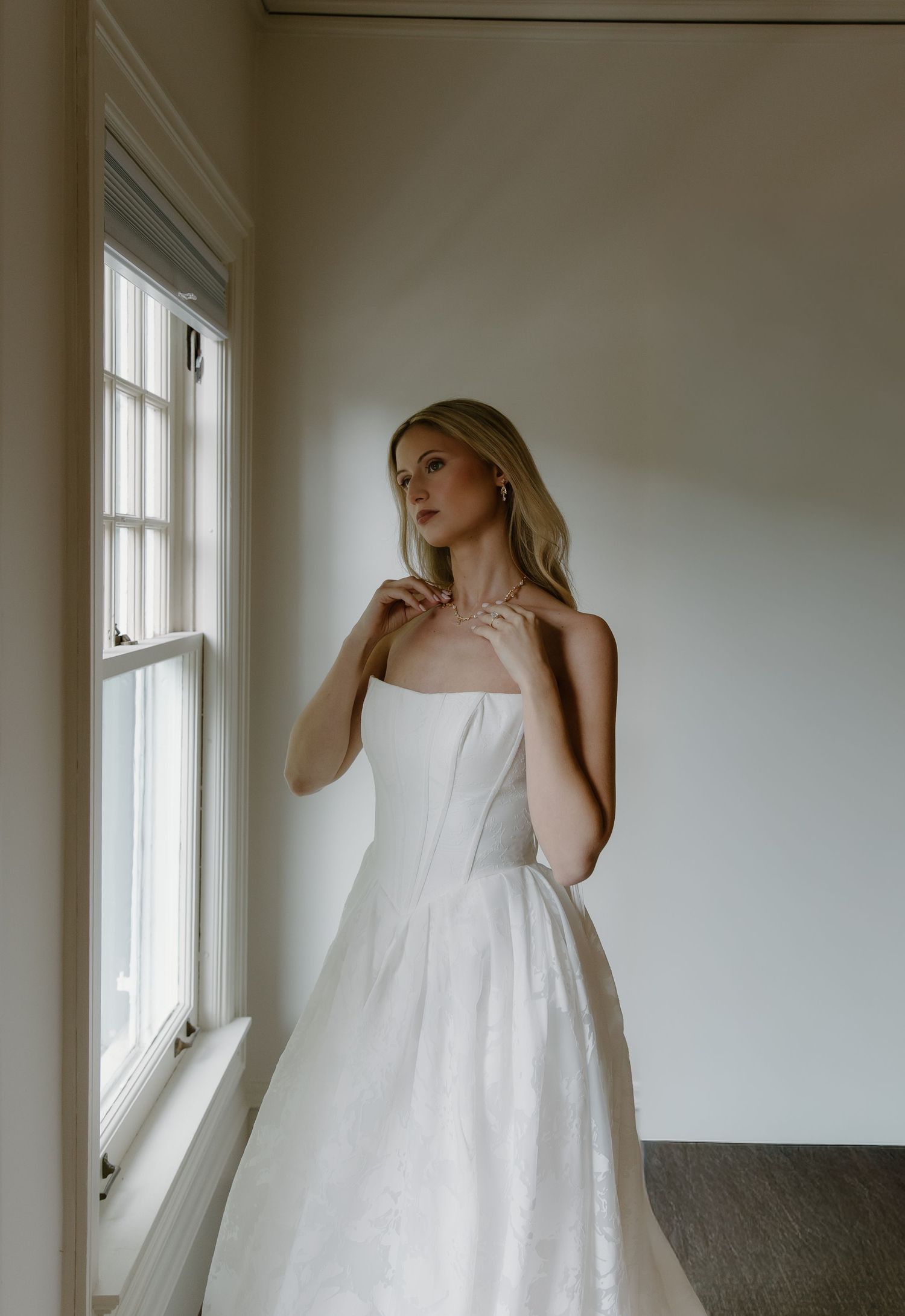 Elegant bride in a strapless white ball gown standing by a sunlit window in a minimalist room.
