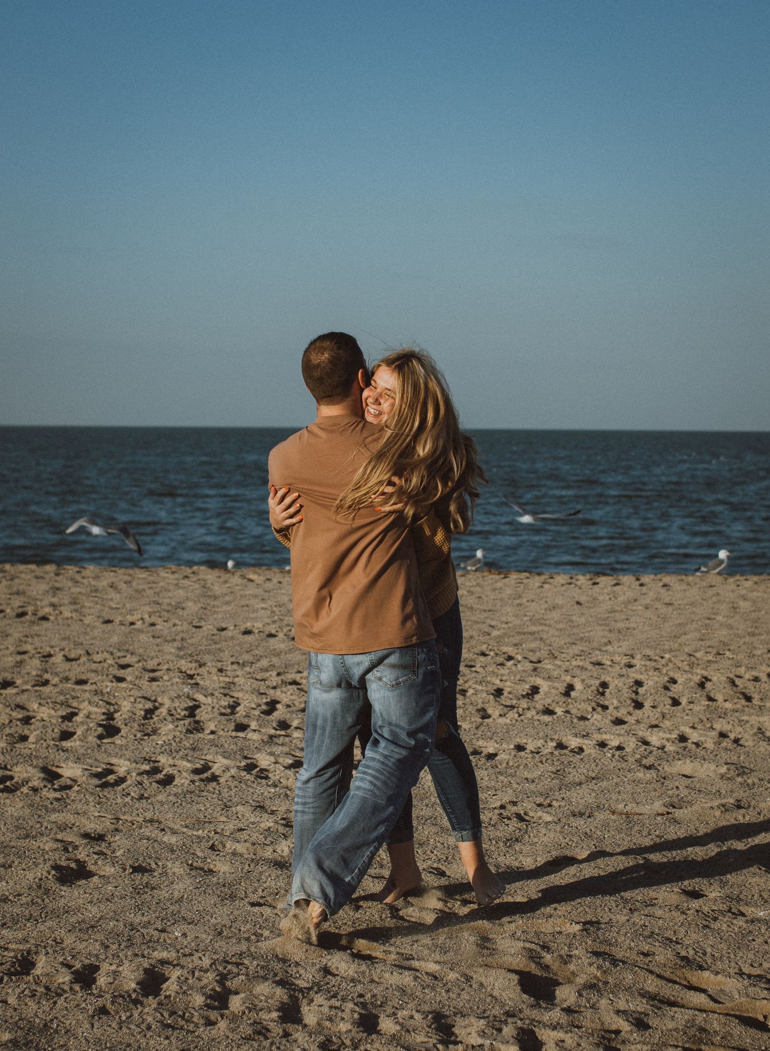 Couple embracing while standing barefoot on a sandy beach with ocean views.
