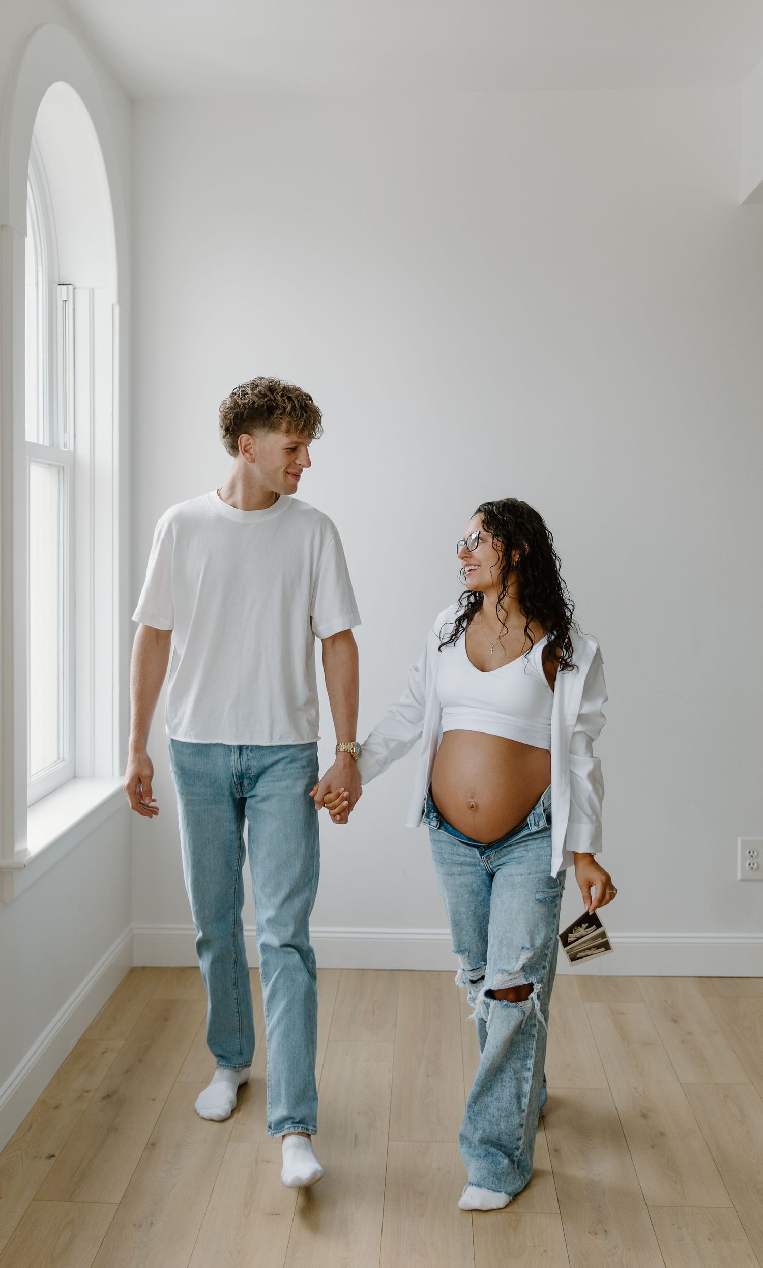 A pregnant couple walks hand-in-hand in a bright white room, smiling at each other, wearing casual jeans and white tops.