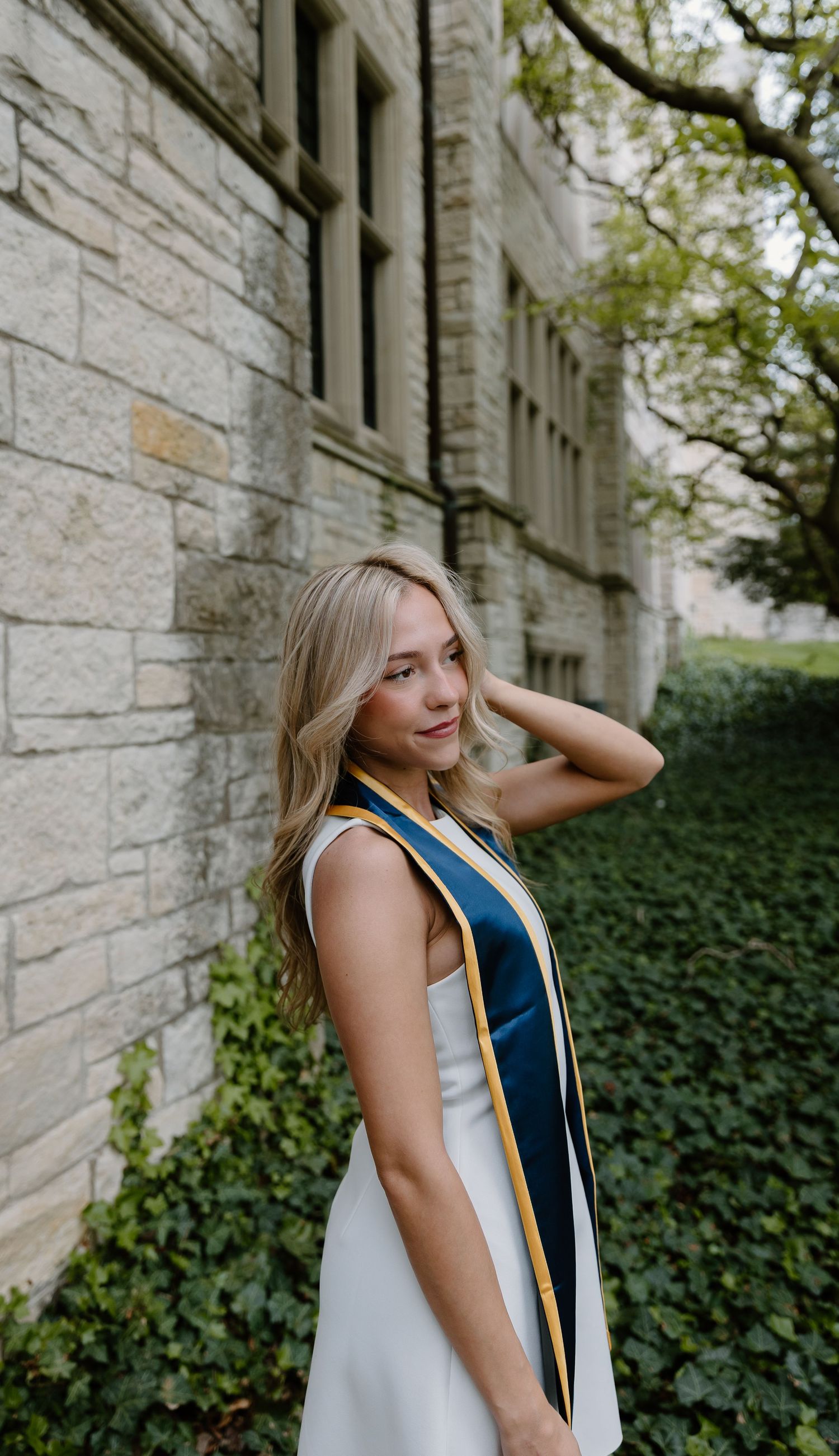 Blonde woman in white dress and navy graduation stole poses outdoors near stone building wall with ivy.