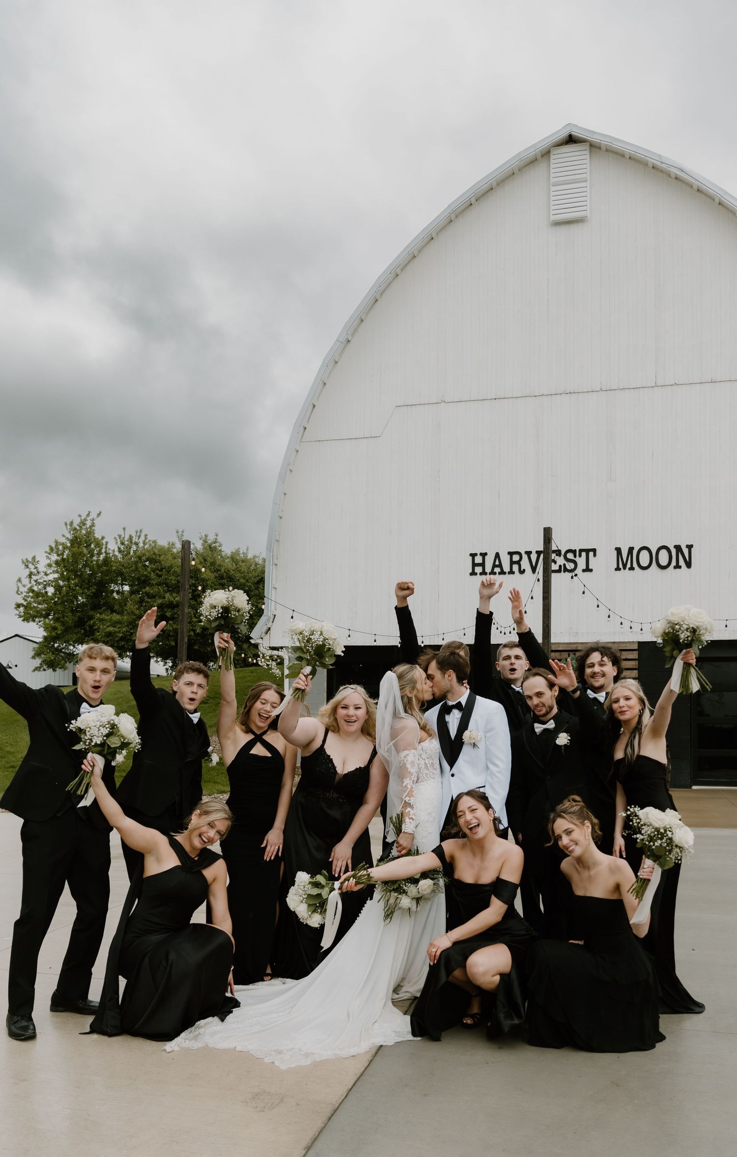 Joyful wedding party celebrates in front of white Harvest Moon barn with bridesmaids in black gowns.