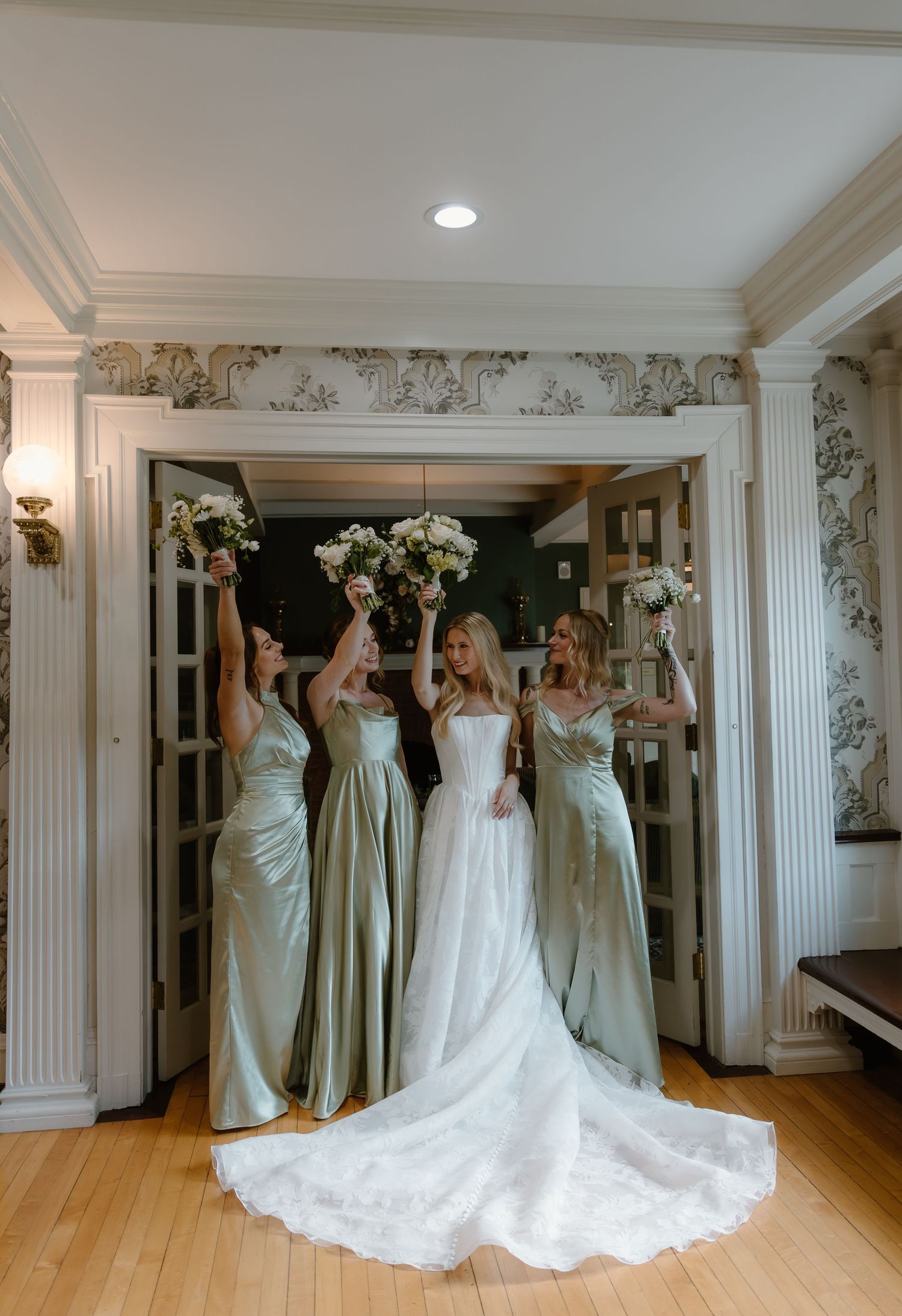 Bride with three bridesmaids in sage green satin gowns raising bouquets inside an elegant venue.