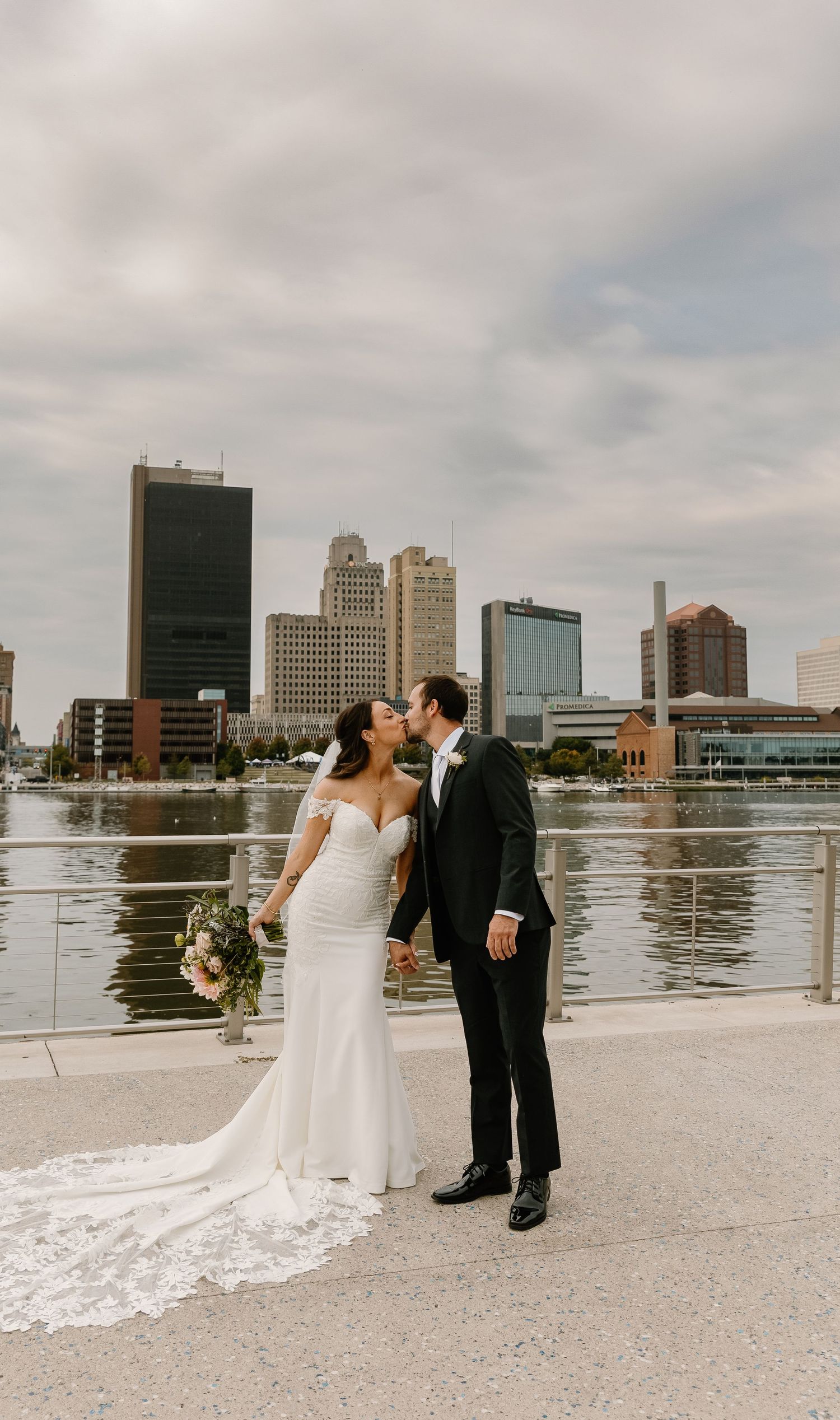 A romantic wedding couple shares a kiss on a waterfront terrace with the Toledo city skyline in the background.
