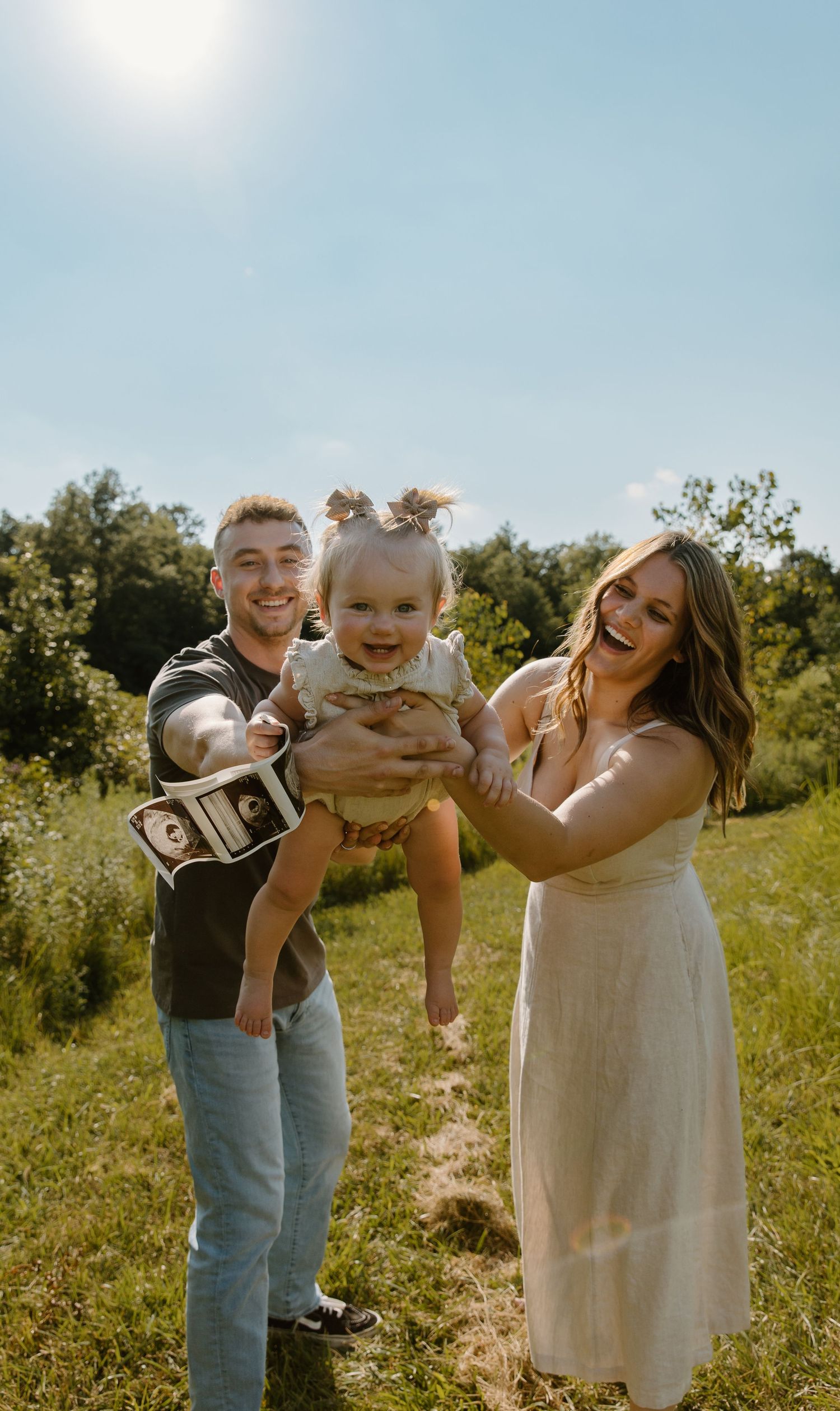 Family plays together outdoors in a sunny field holding up ultrasound photos and celebrating.