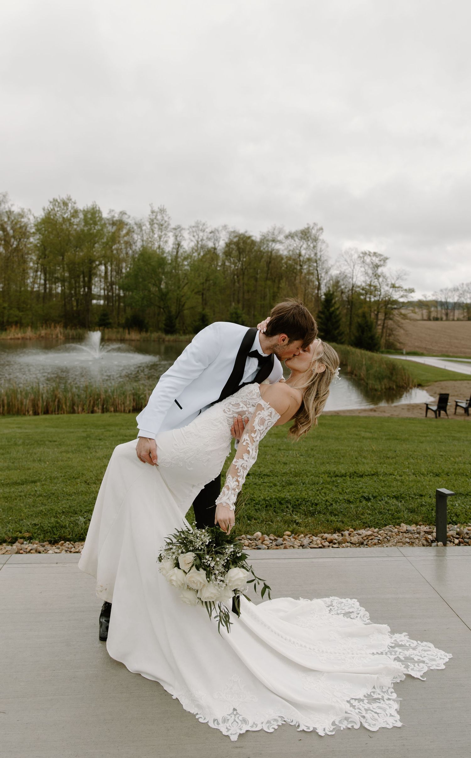Bride and groom share a romantic dip kiss on patio with scenic pond and green trees backdrop.