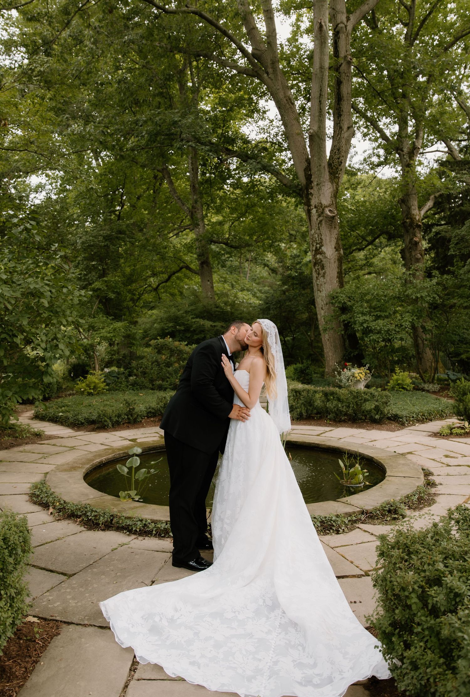 Newlyweds share a romantic kiss beside a circular garden pond surrounded by lush greenery and stone pathways.