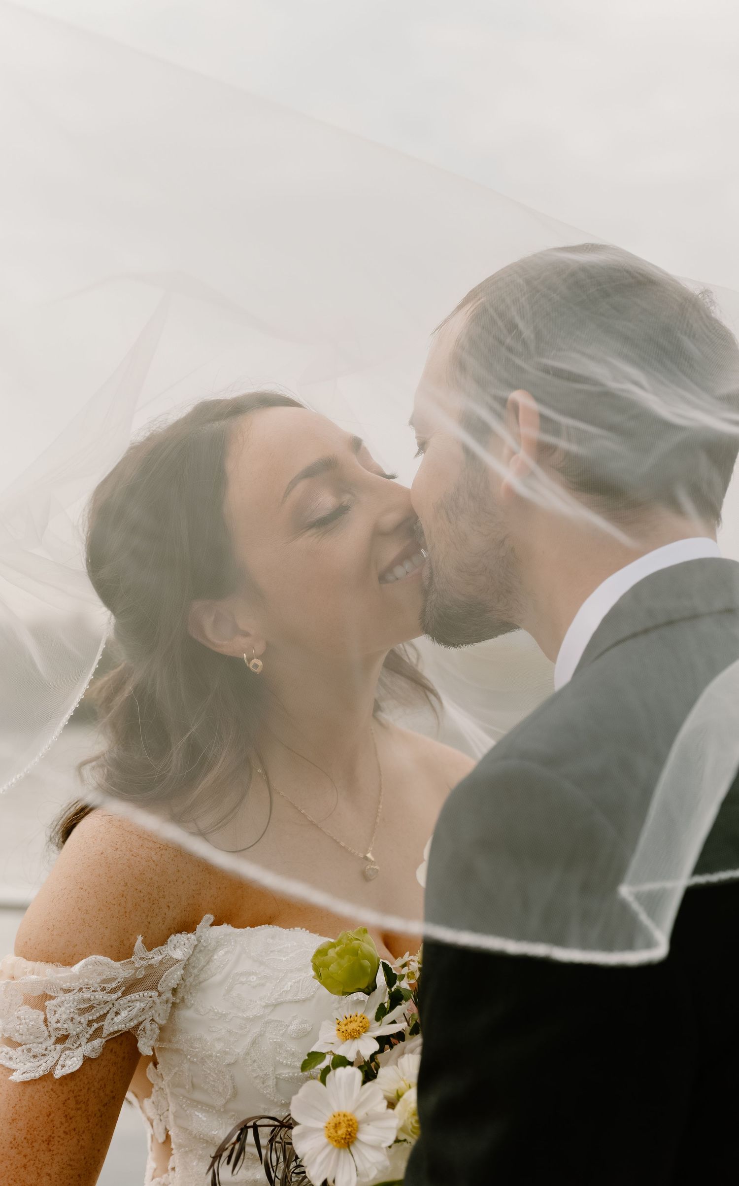 A romantic wedding couple shares a tender kiss under a flowing veil with delicate floral details.