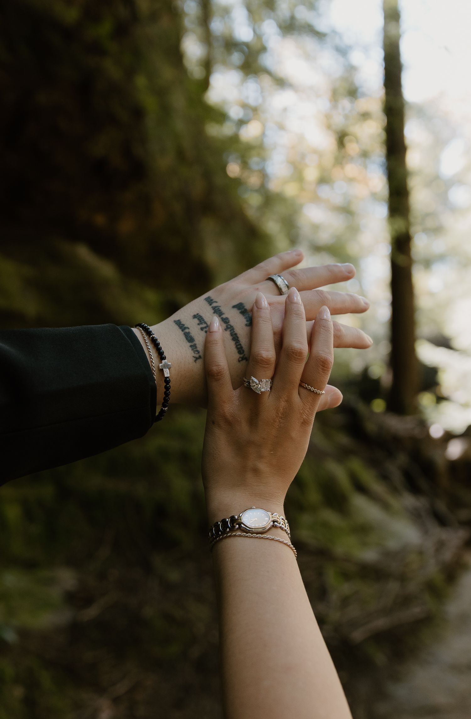 Two hands touching in a forest, one with tattoos and rings, the other wearing a delicate bracelet and watch.