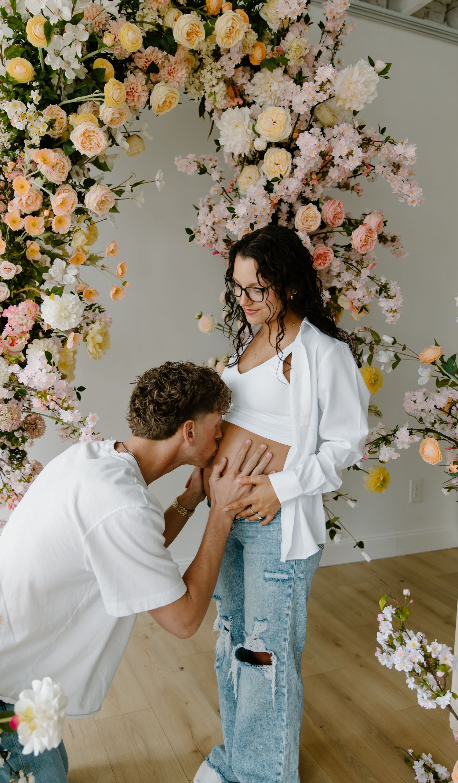 A man kisses a pregnant woman's belly in front of a floral arch with peach and pink blooms.
