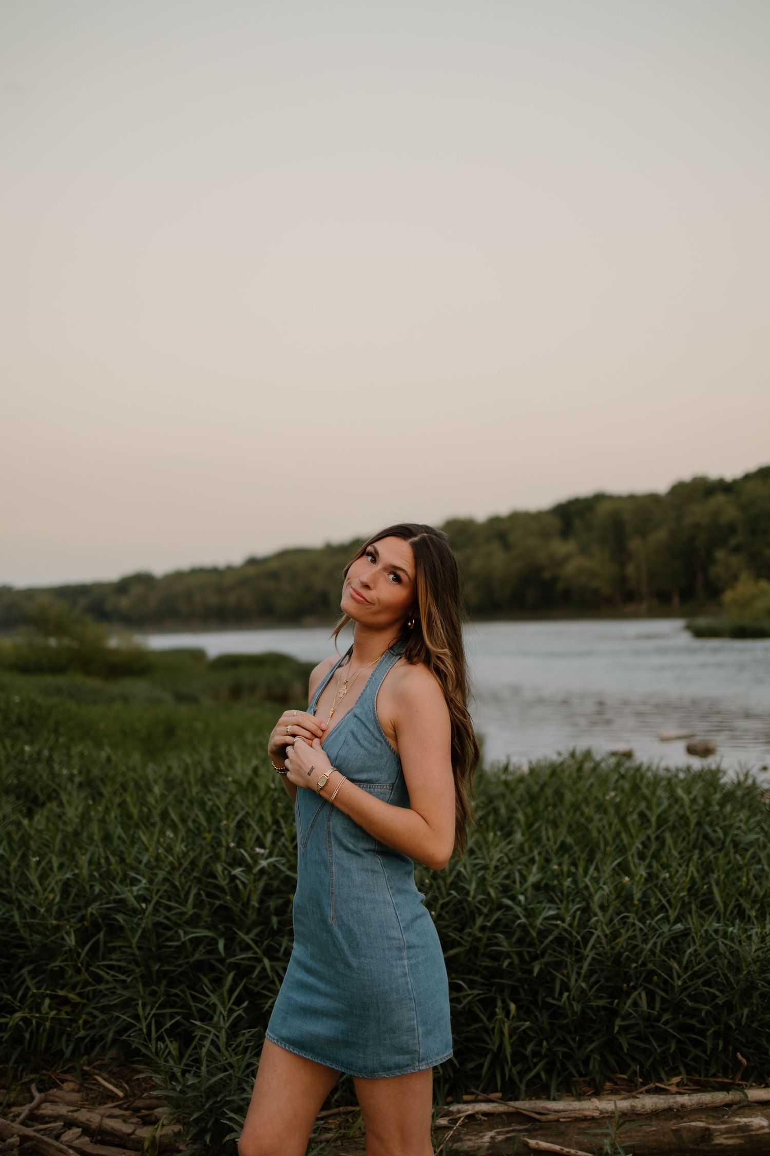 Brunette woman in blue mini dress posing by lush green riverbank during golden hour.