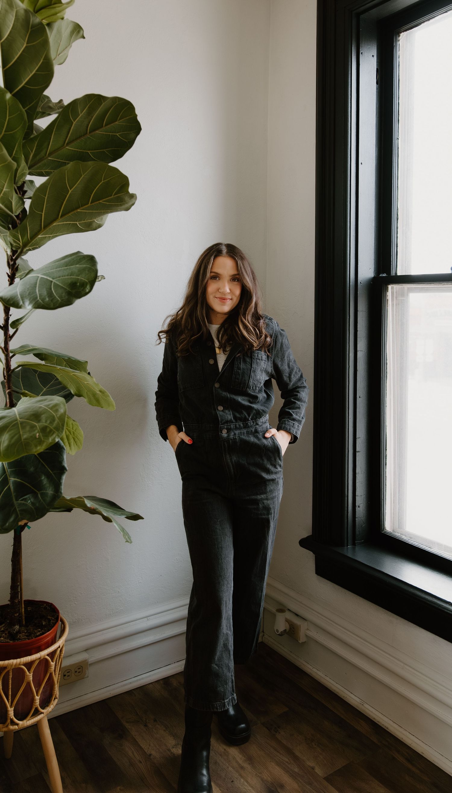 Woman in black denim jumpsuit posing indoors near a fiddle leaf fig plant and black-framed window.