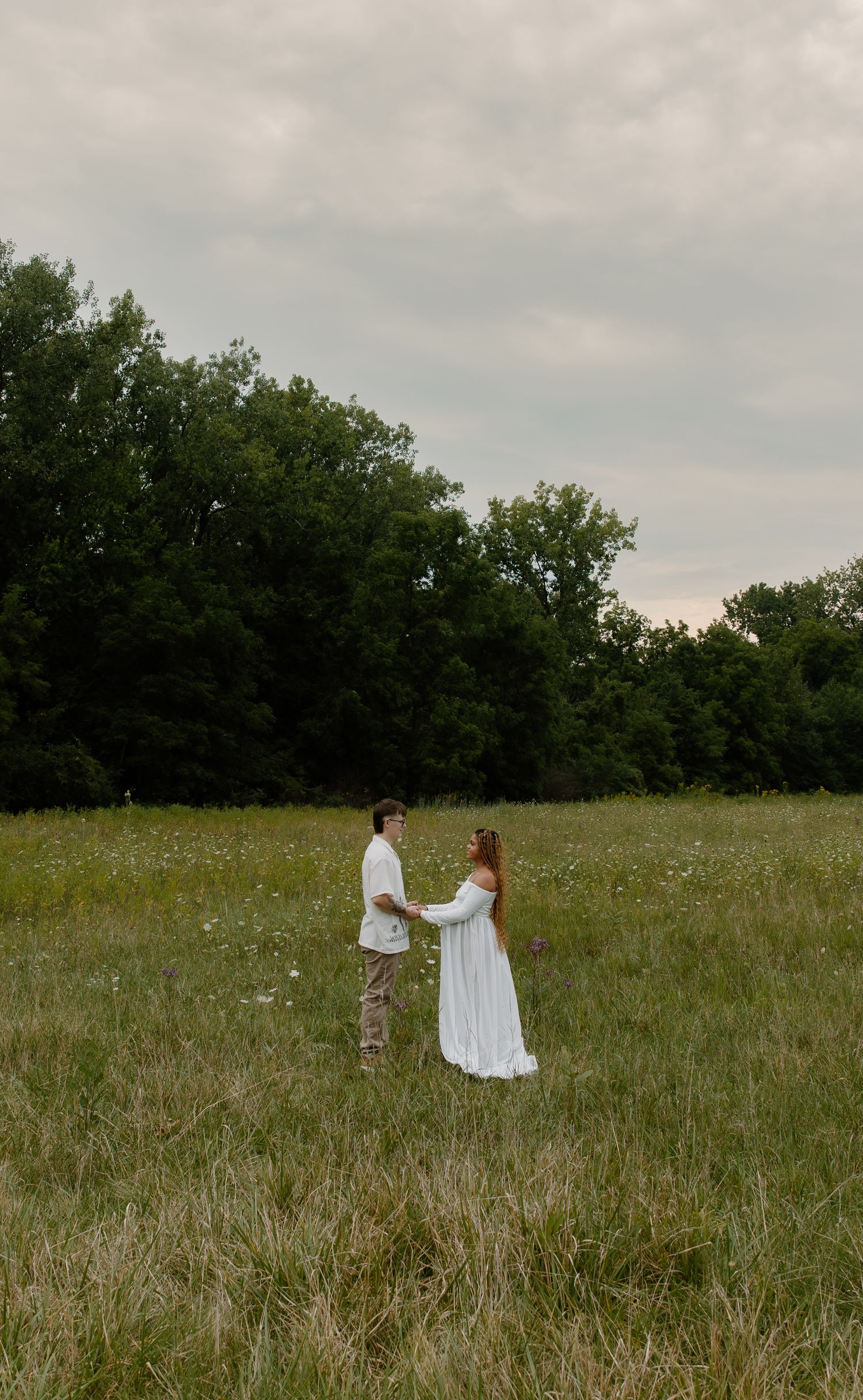 A couple in white attire holds hands in a lush green meadow with wildflowers, trees, and a cloudy sky backdrop.