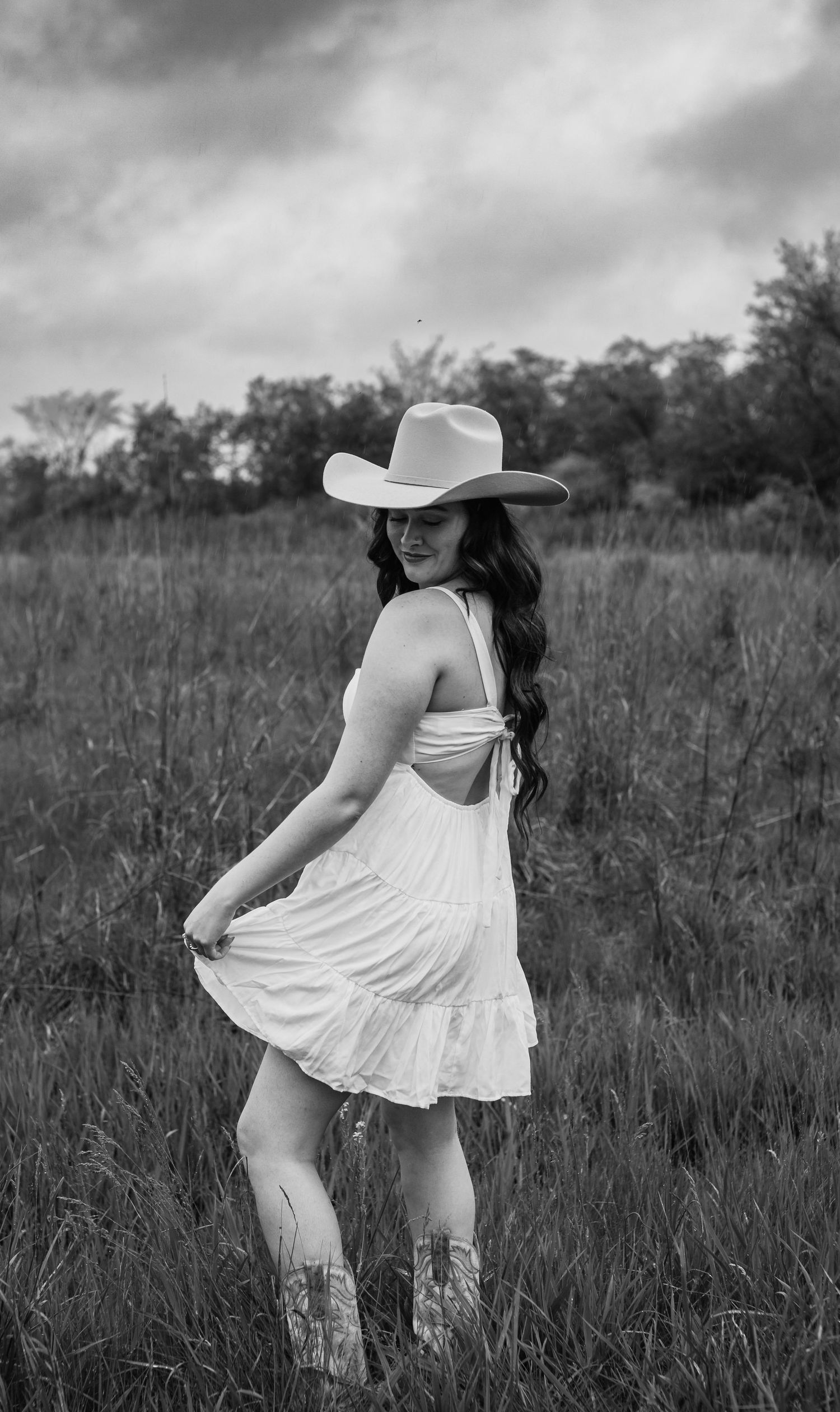 Black and white photo of a figure in a white sundress and cowboy hat twirling in a field of tall grass.