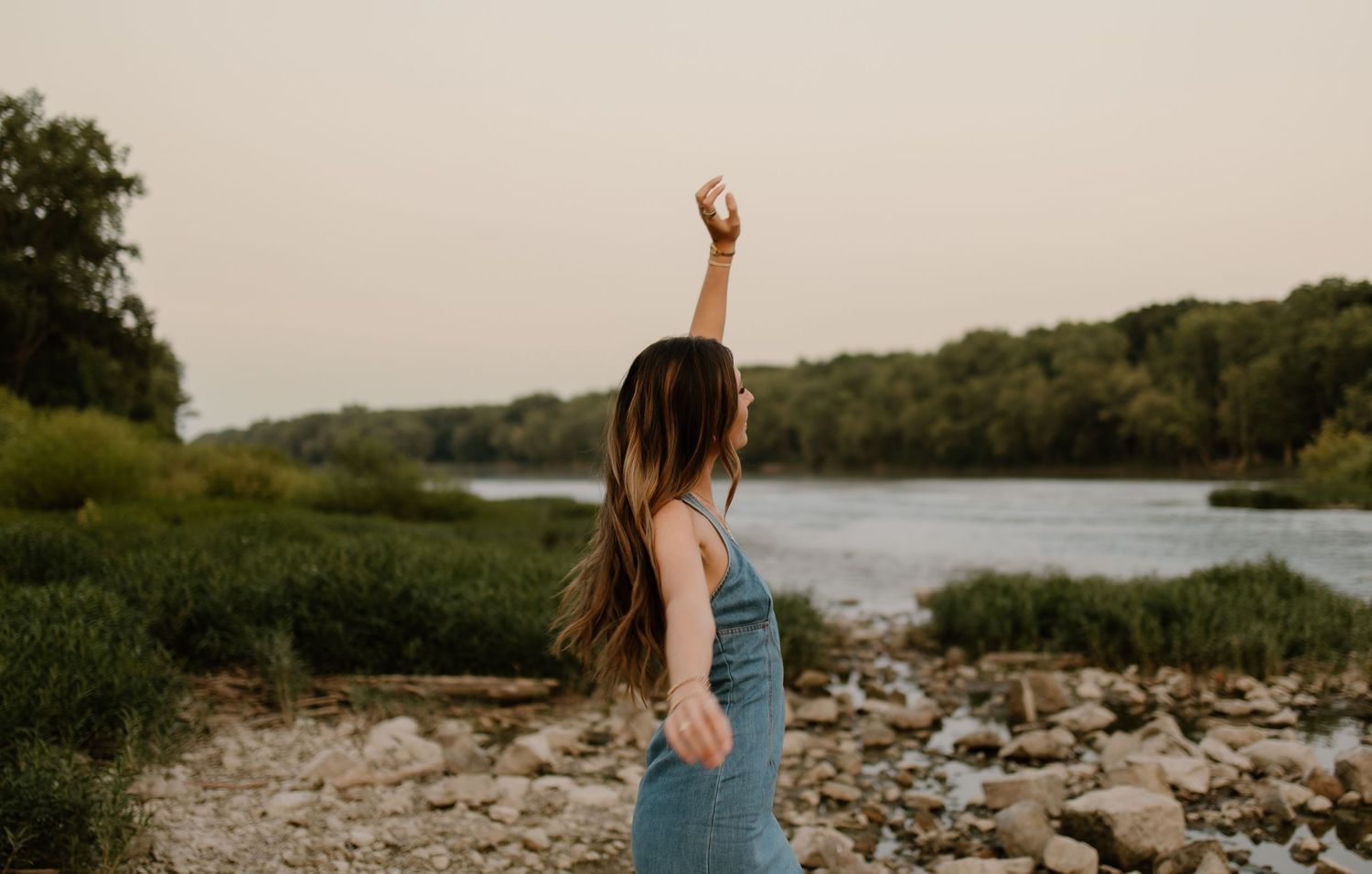 A young woman in a blue dress raises her arm by a rocky riverbank surrounded by lush green trees at dusk.