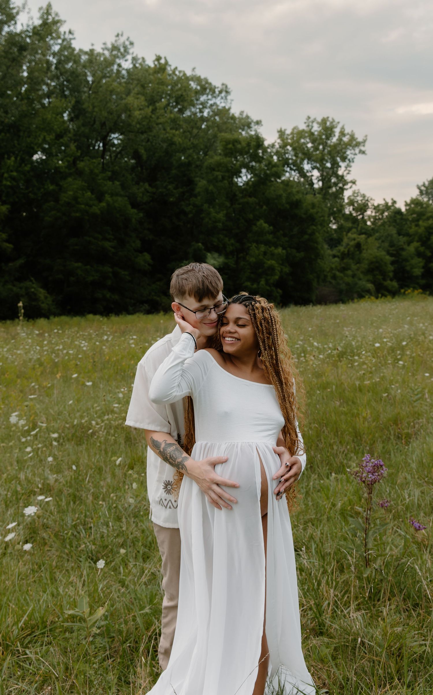 Couple in a wildflower field, woman in white maternity gown with floral crown, partner embracing her from behind.