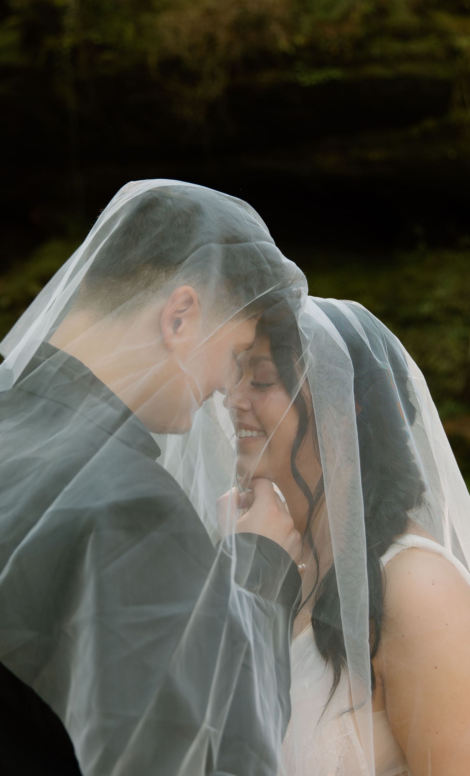 A bride and groom share an intimate moment under a flowing white wedding veil outdoors.
