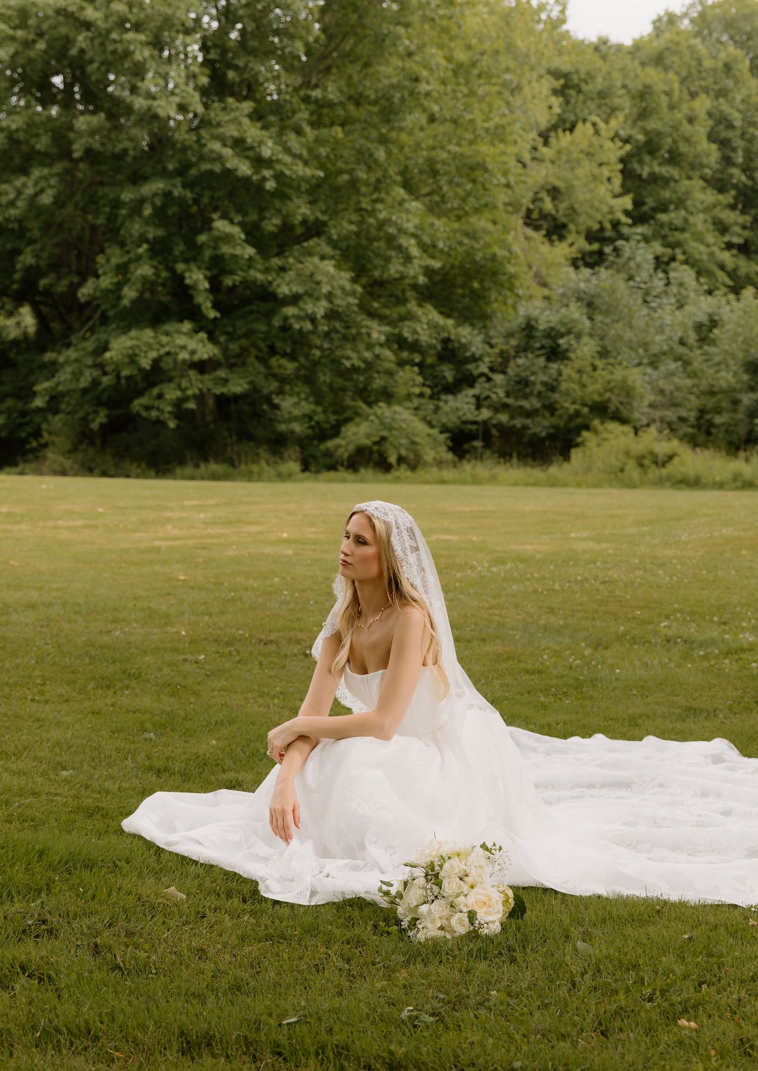 Bride in flowing white gown and veil sits gracefully on green lawn with bouquet, surrounded by lush trees.