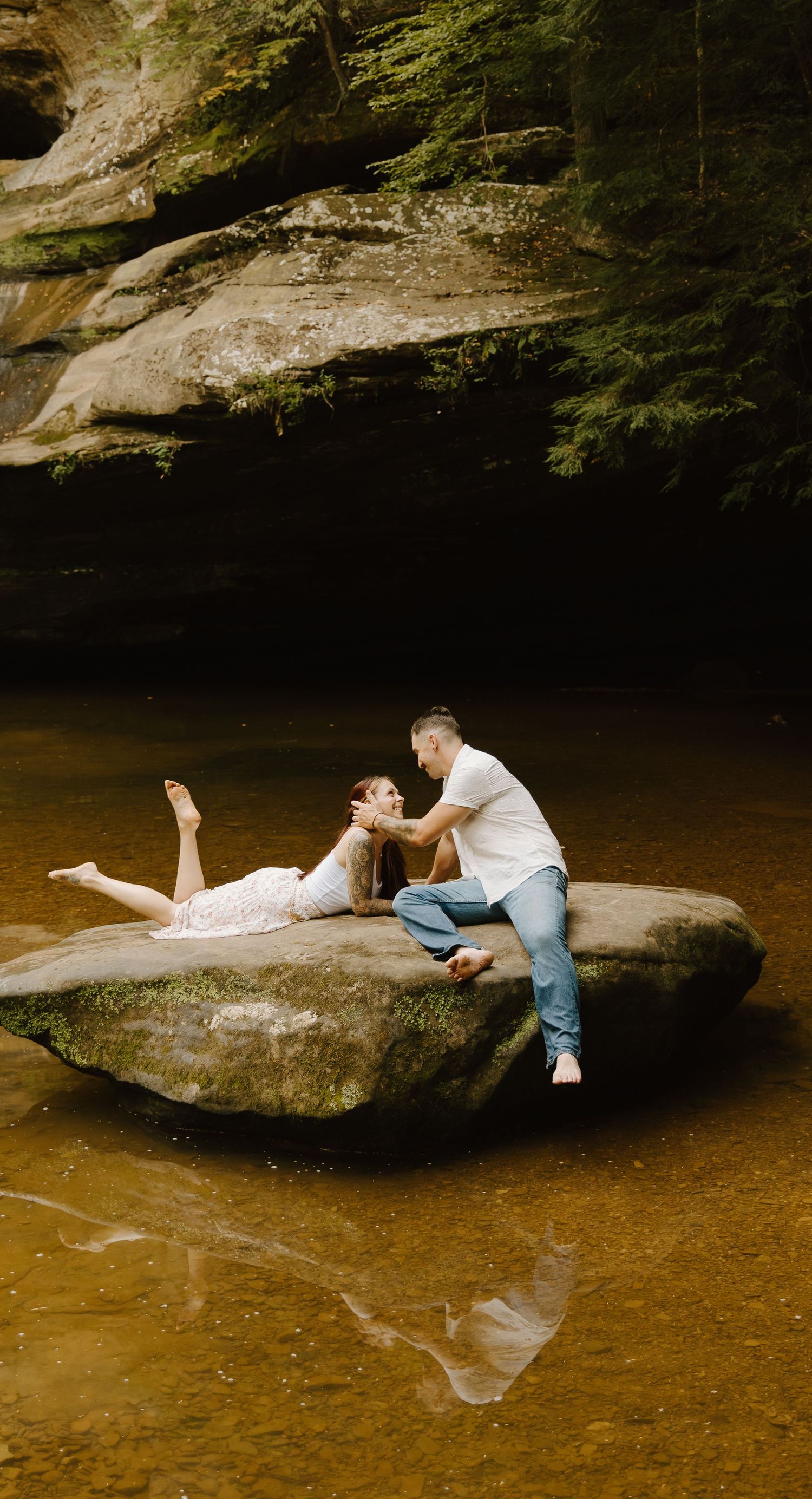 Couple sharing a romantic moment on a large rock in a serene forest stream, woman in white dress, man in white shirt.