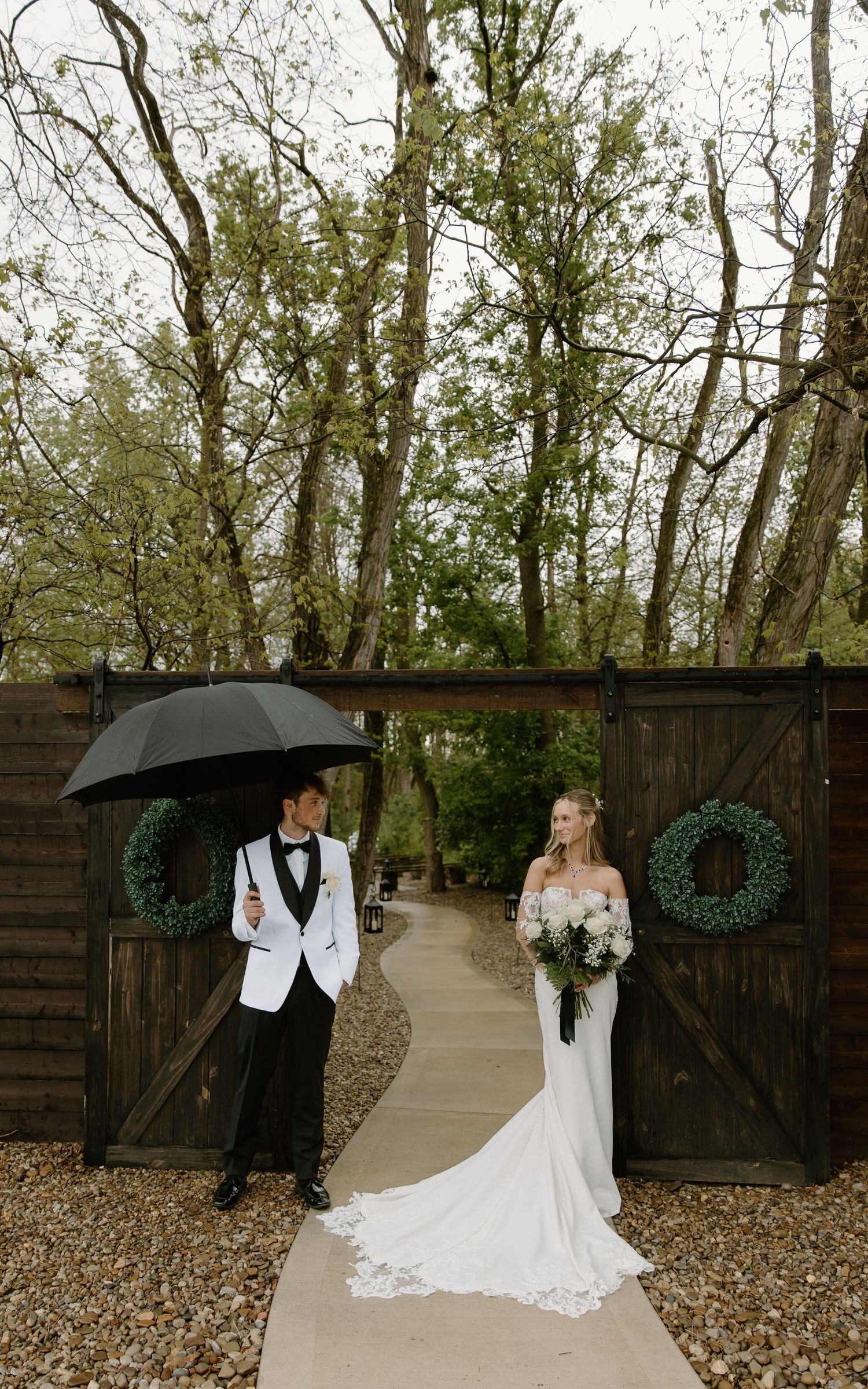Bride in white gown and groom in white tuxedo pose by rustic barn doors with wreaths on a rainy wedding day.
