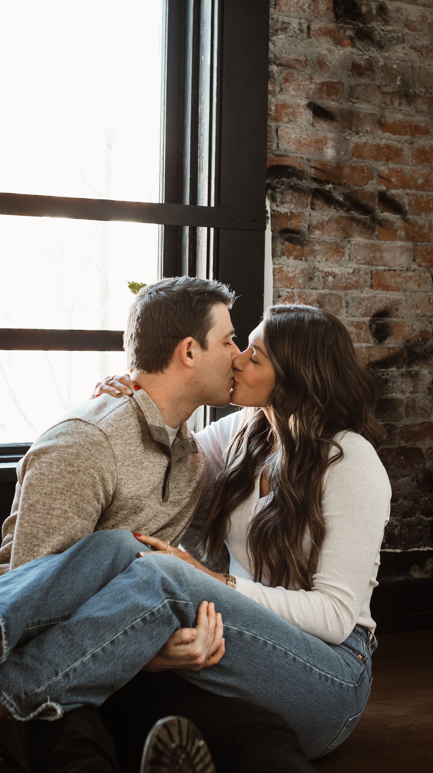 Couple kissing by a rustic brick wall window in casual attire.