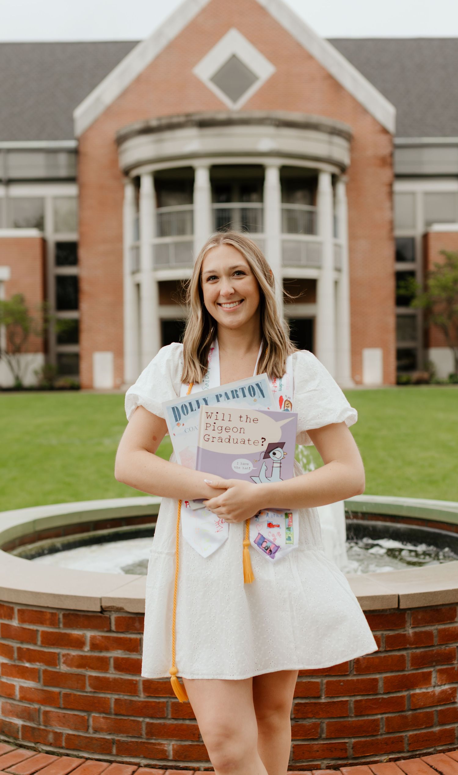 A person stands smiling outside a brick building with a curved architectural feature and circular fountain.