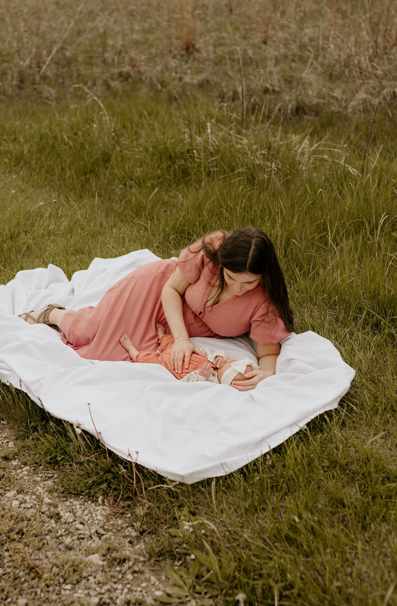 Someone in a peach-colored dress lies on a white blanket in a grassy field reading a book on a sunny day.