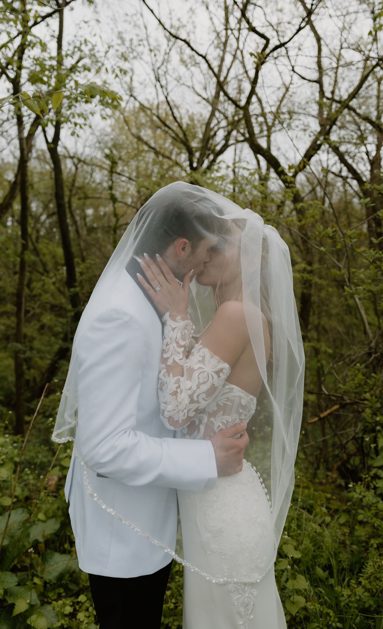 Newlywed couple sharing a romantic kiss under a flowing bridal veil outdoors, surrounded by lush green trees.