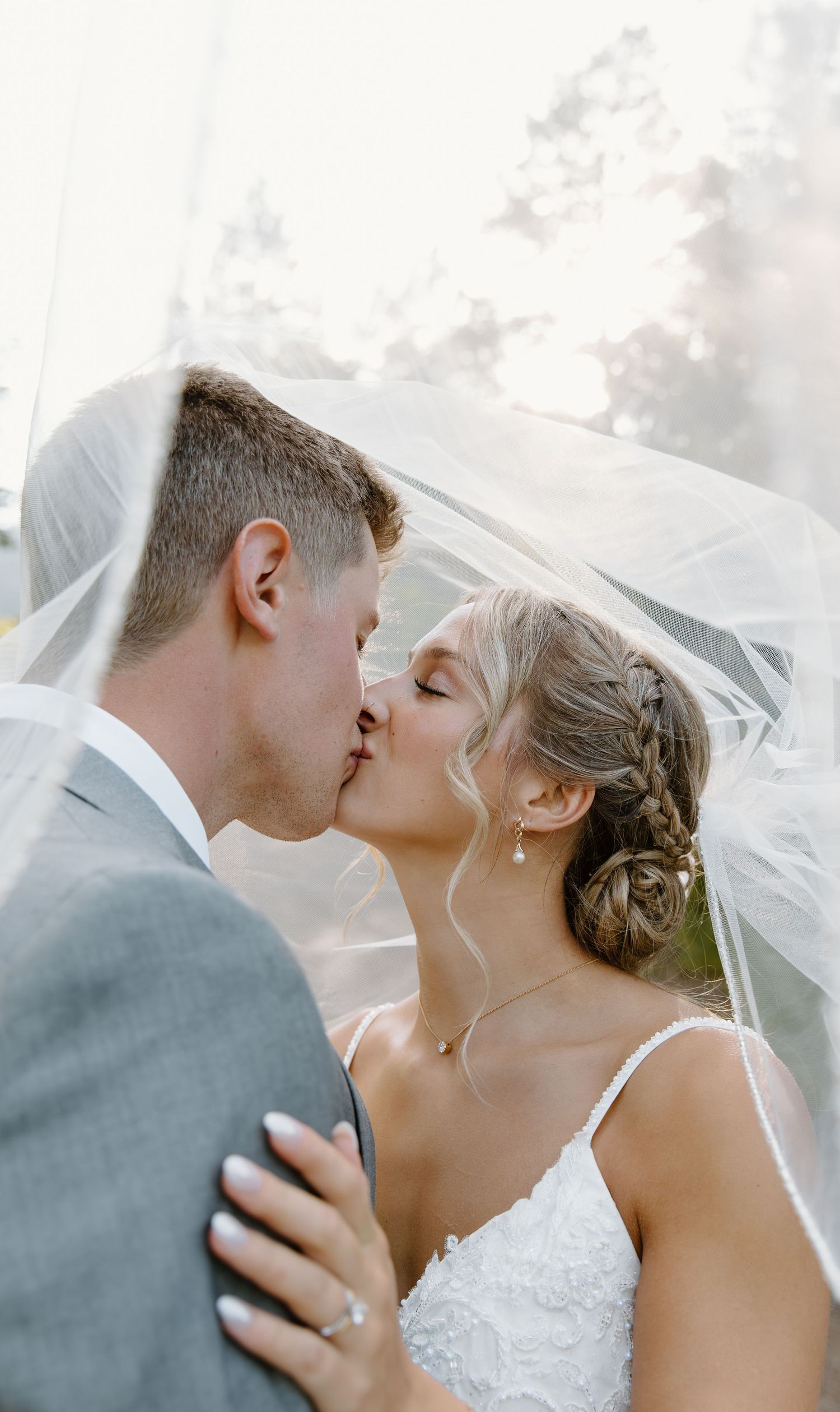 Romantic wedding couple sharing intimate kisses under flowing white veil during outdoor photo session.